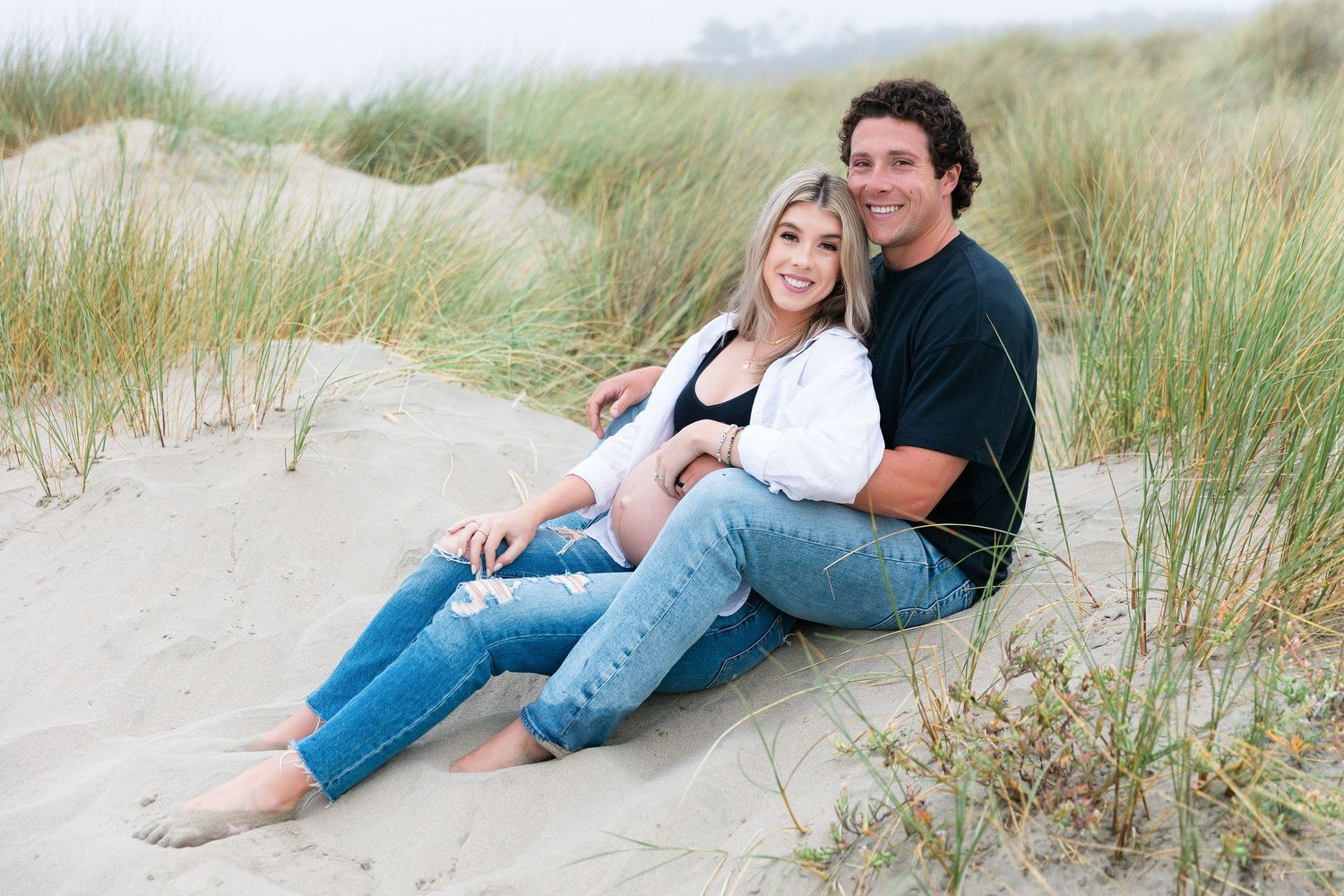 Couple relaxing together on sandy beach dunes surrounded by coastal grass.