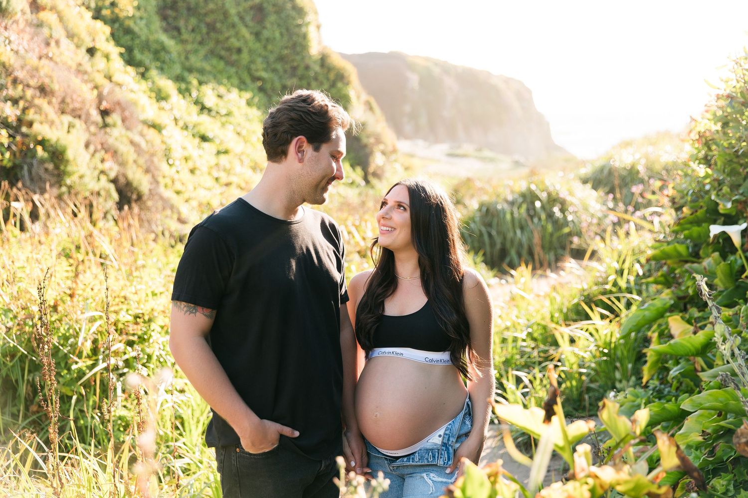 Expecting couple smiles together during golden hour maternity session among wildflowers.