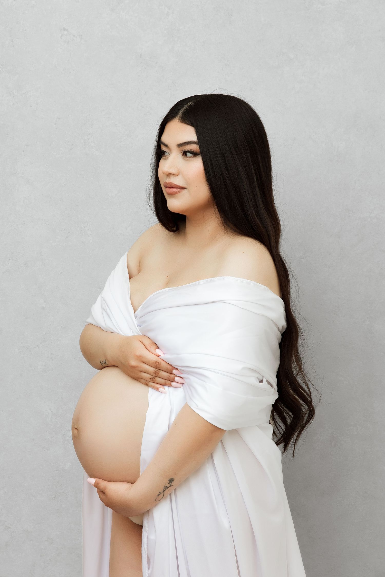 Elegant maternity pose in white off-shoulder gown against grey background.