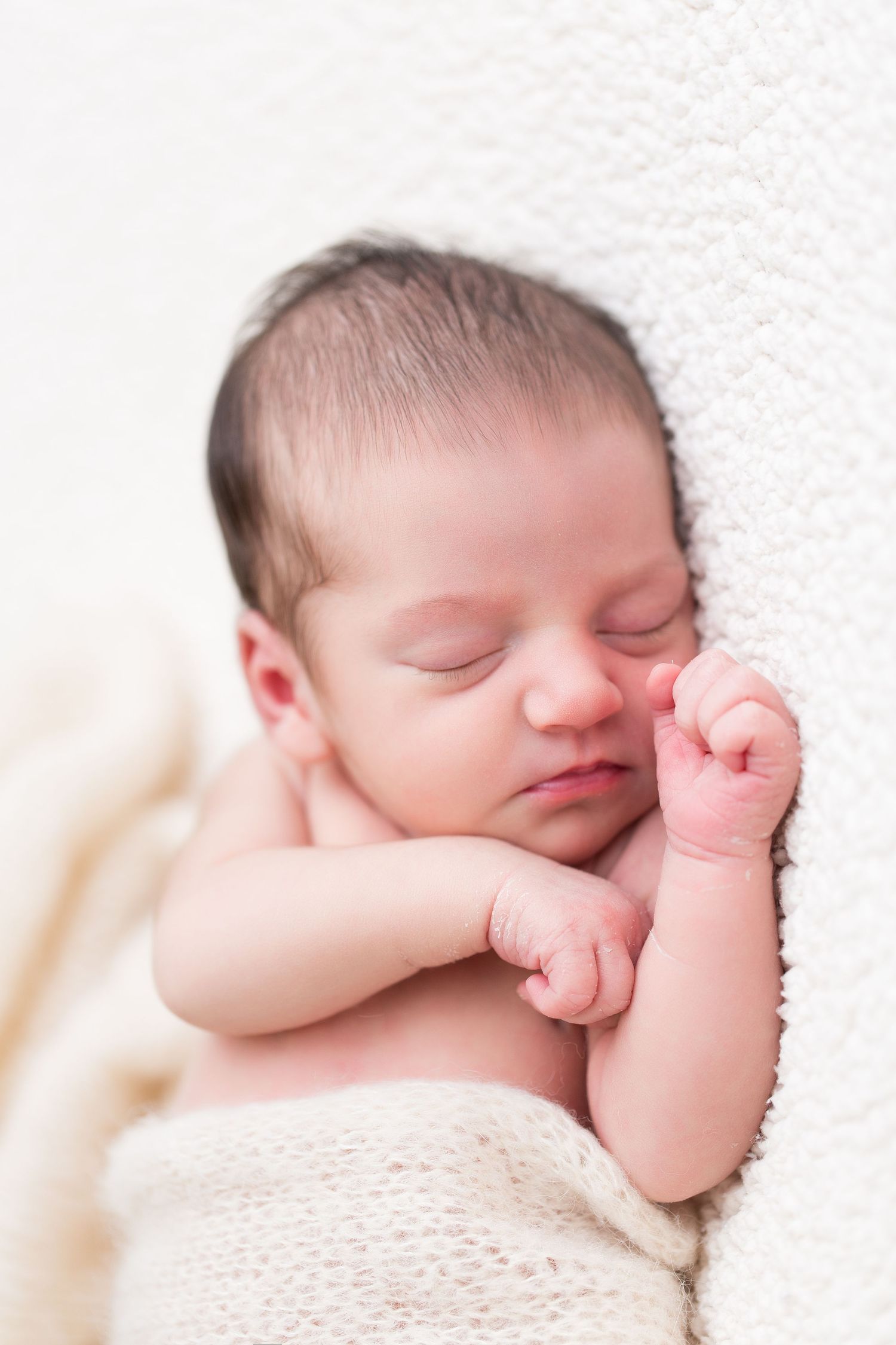 Peaceful sleeping newborn baby wrapped in soft white knitted blanket with tiny fist curled up near face.