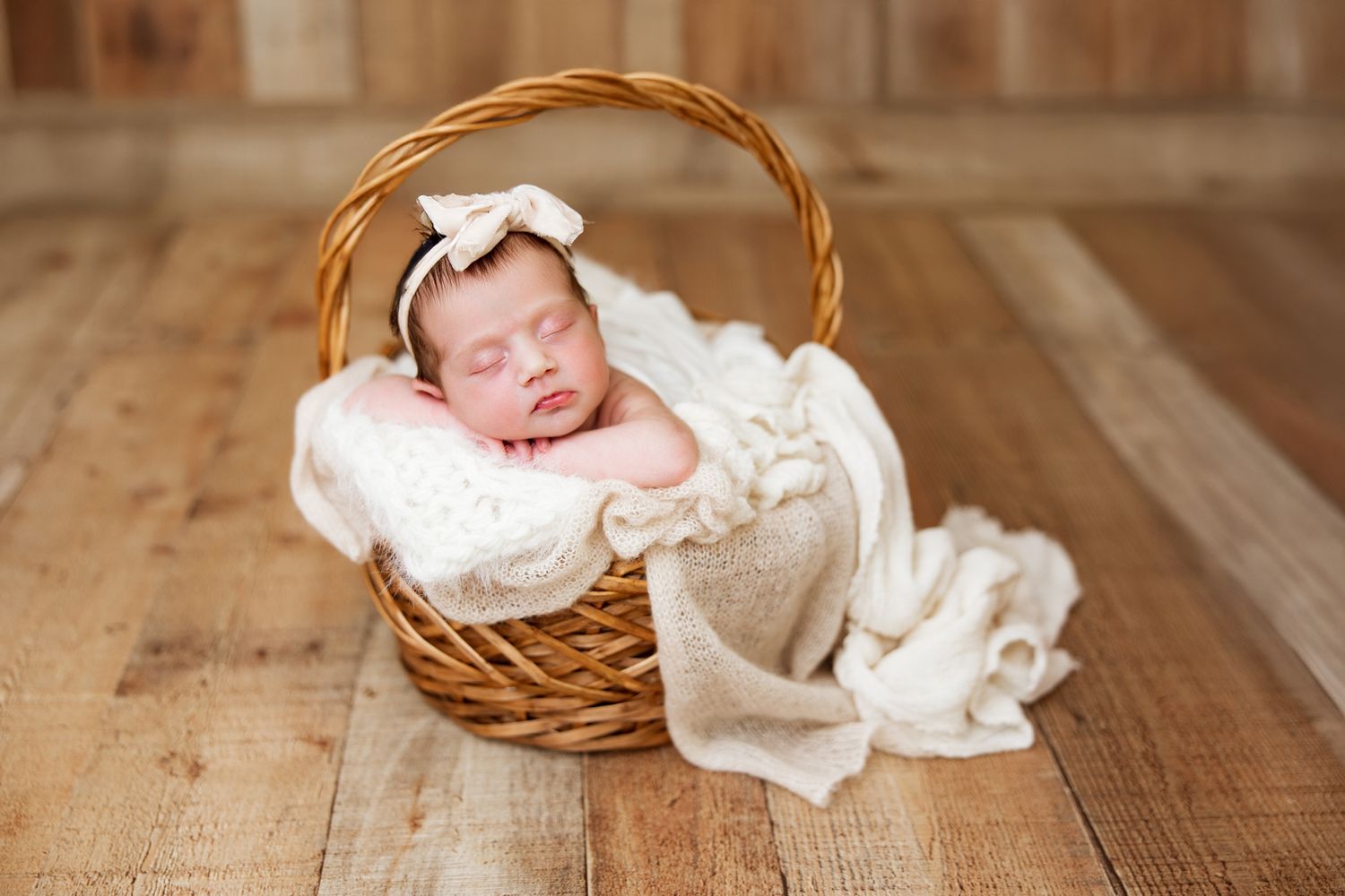 A sleeping newborn baby rests peacefully in a woven basket on a rustic wooden floor.