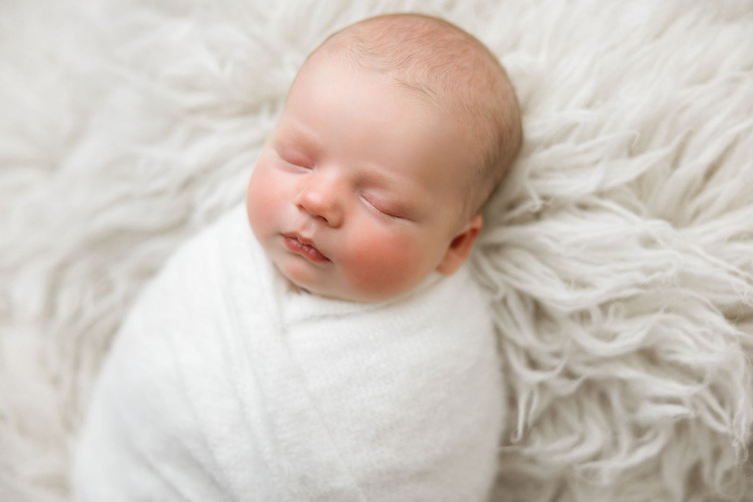 Sleeping newborn baby in white clothing lies peacefully on soft white textured fabric.
