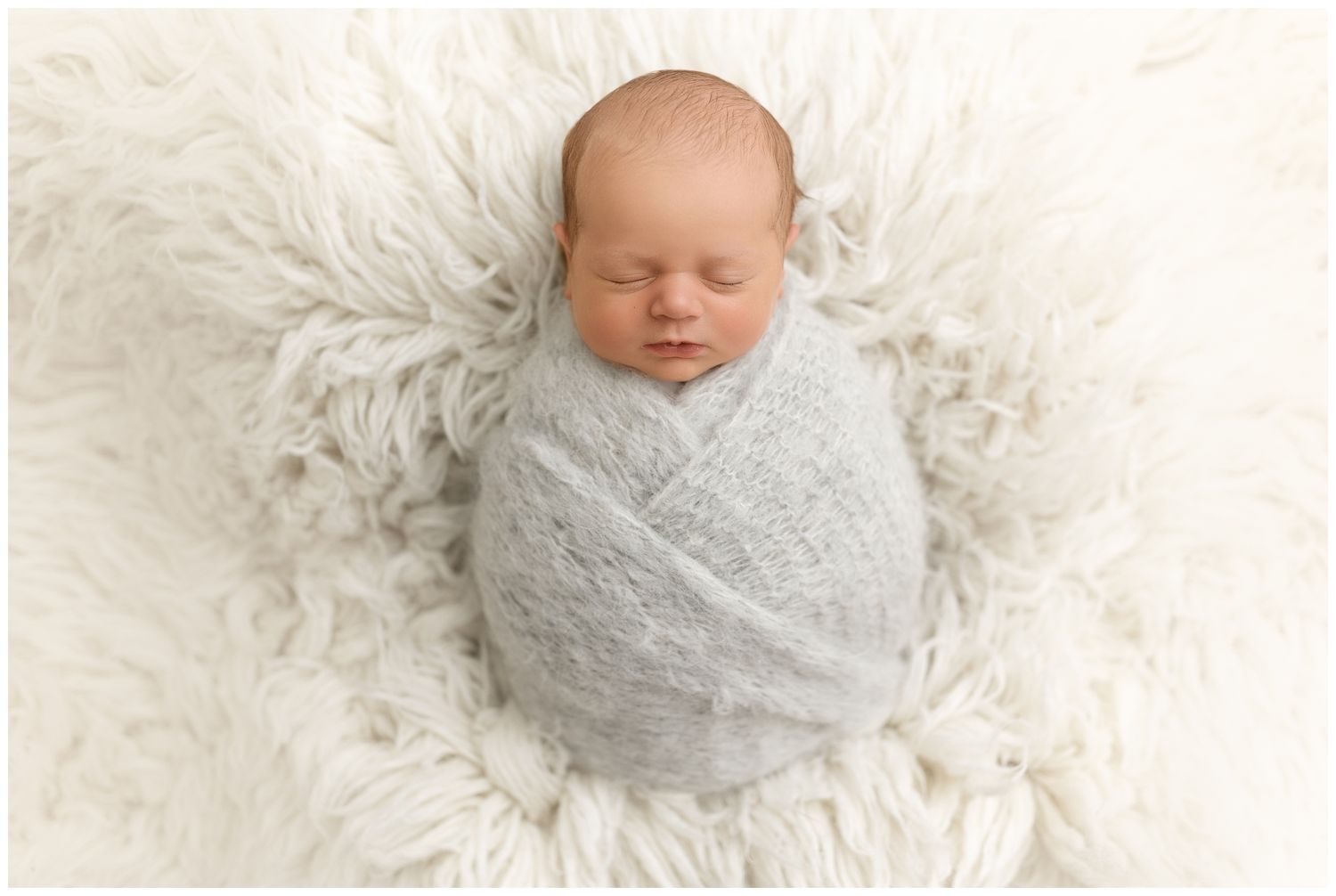 Newborn baby sleeping peacefully wrapped in soft gray swaddle blanket on white fluffy backdrop.