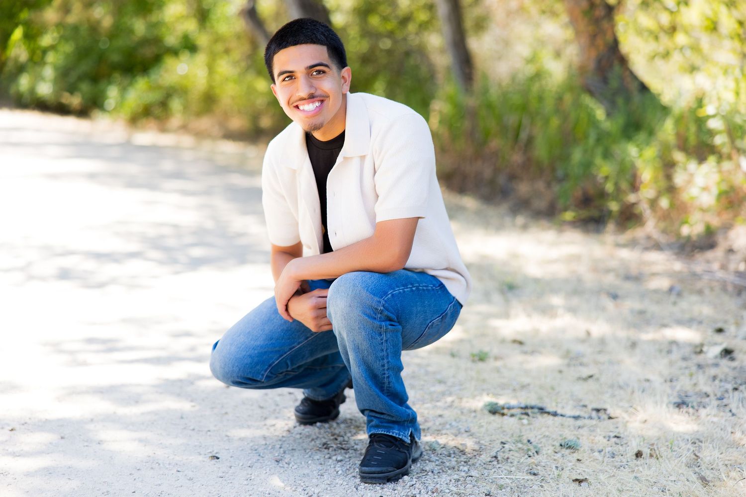 Young man in white shirt and jeans crouching on a dirt path outdoors, smiling confidently.
