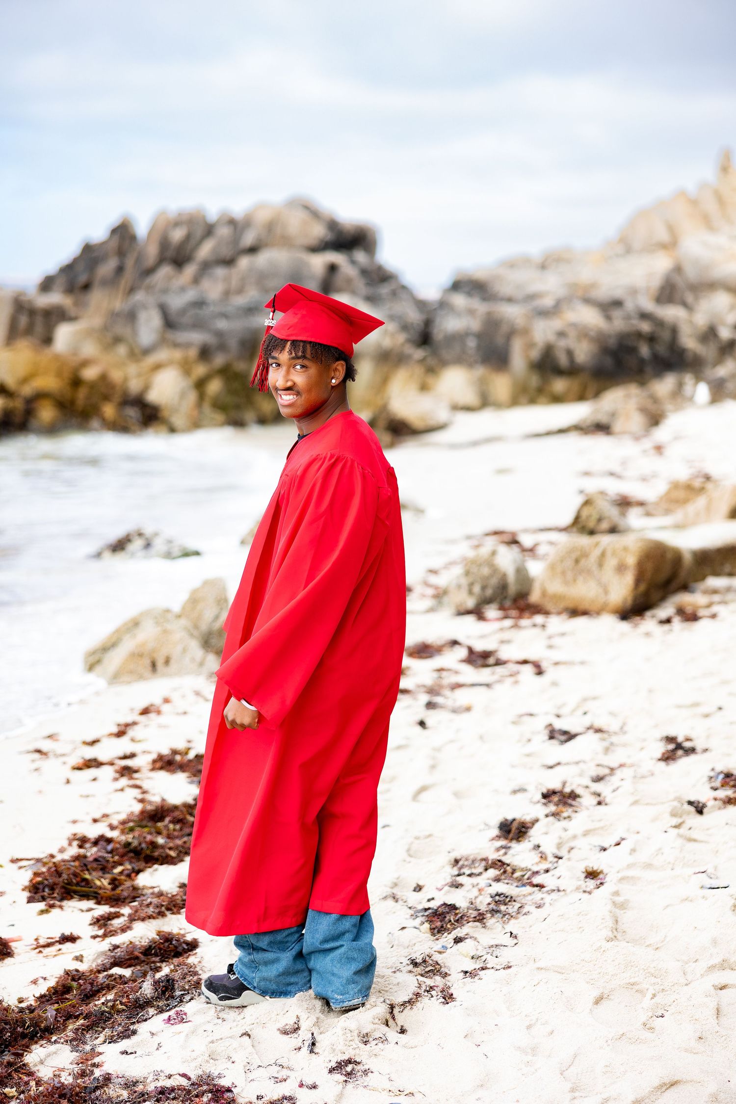 Young man in red graduation cap and gown standing on a rocky beach, smiling at the camera.