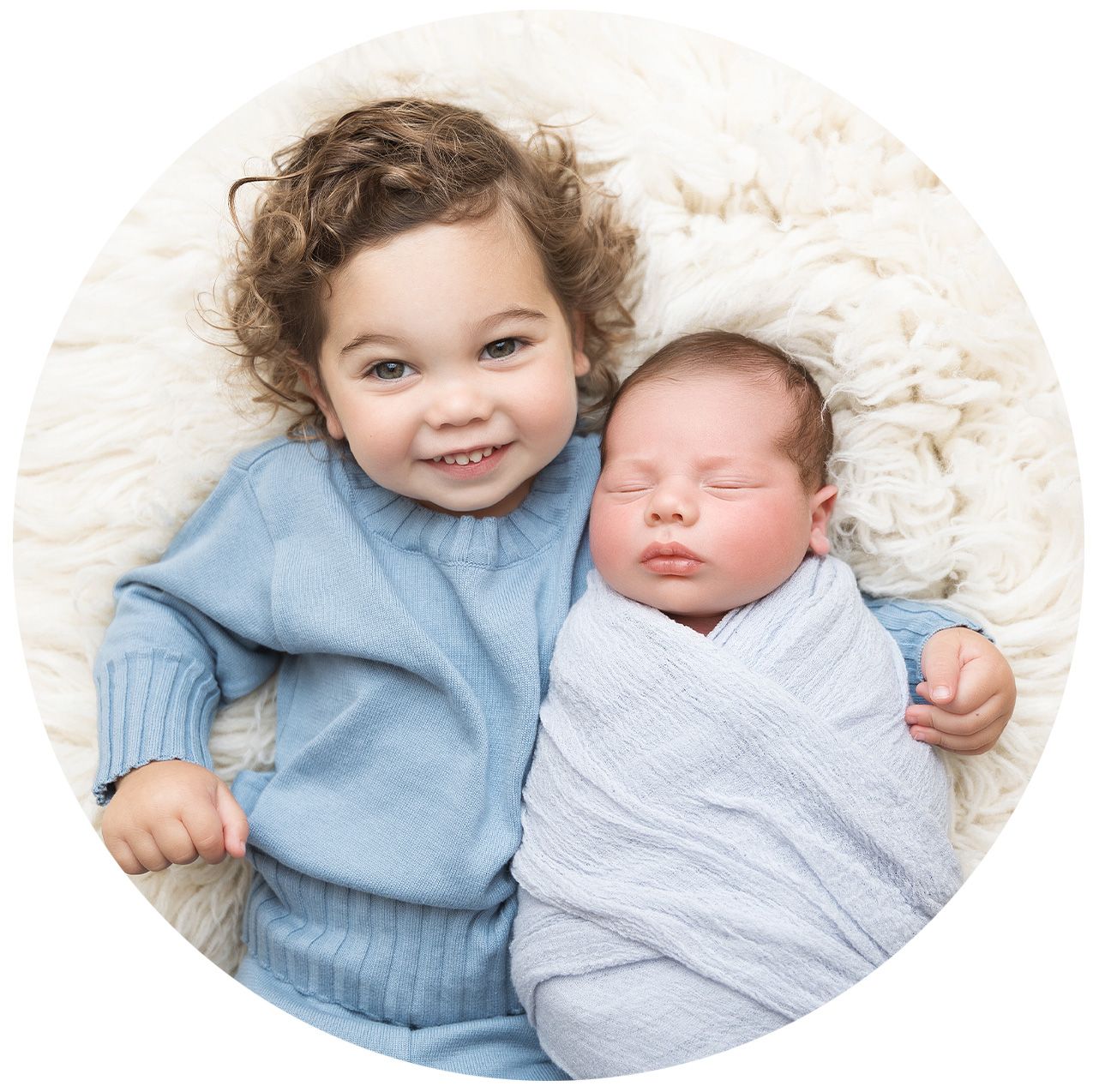 Two young children sit together on a white blanket, with one wearing blue and the other swaddled in light gray.
