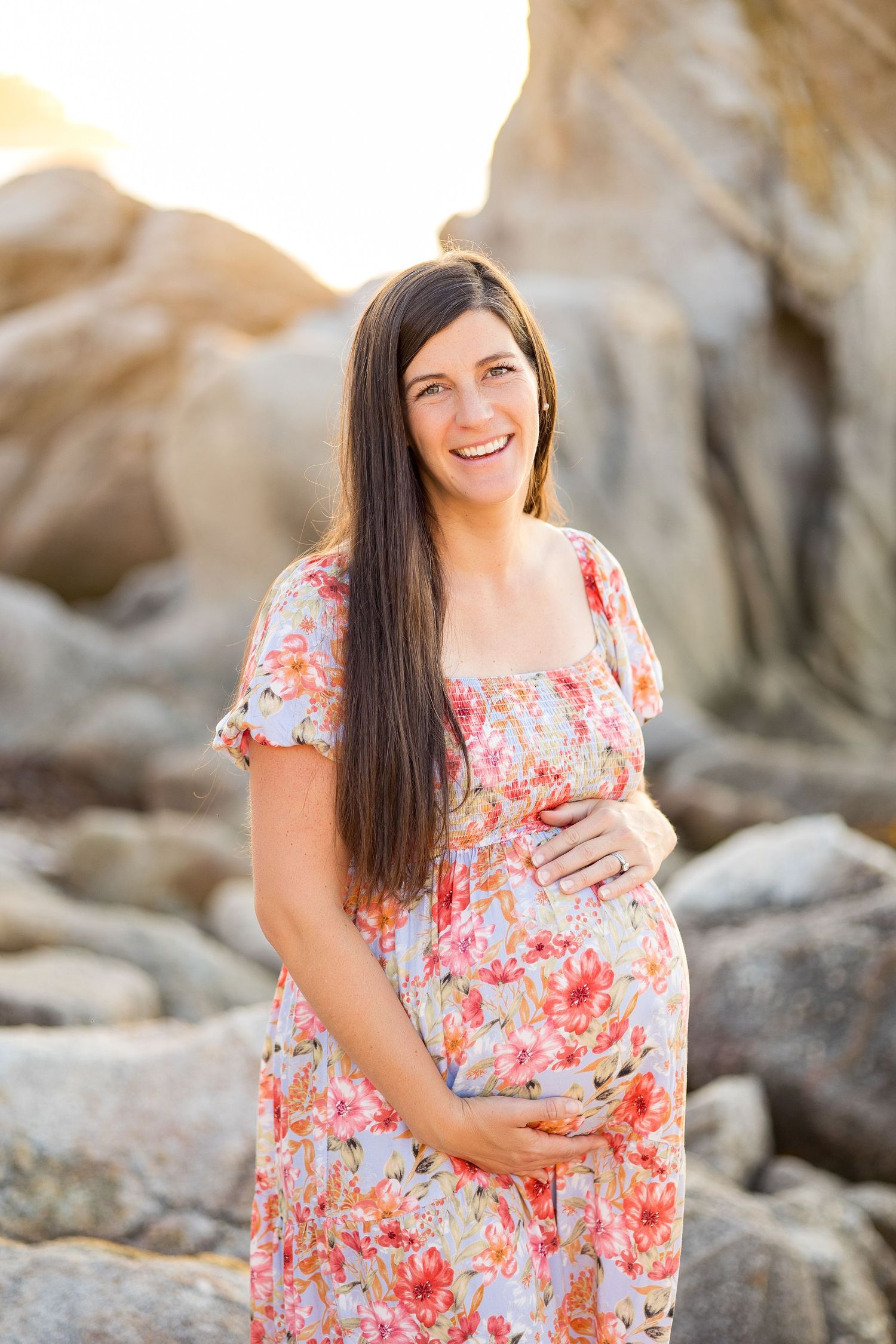 Expectant mother in floral dress poses on rocky beach during golden hour sunset.