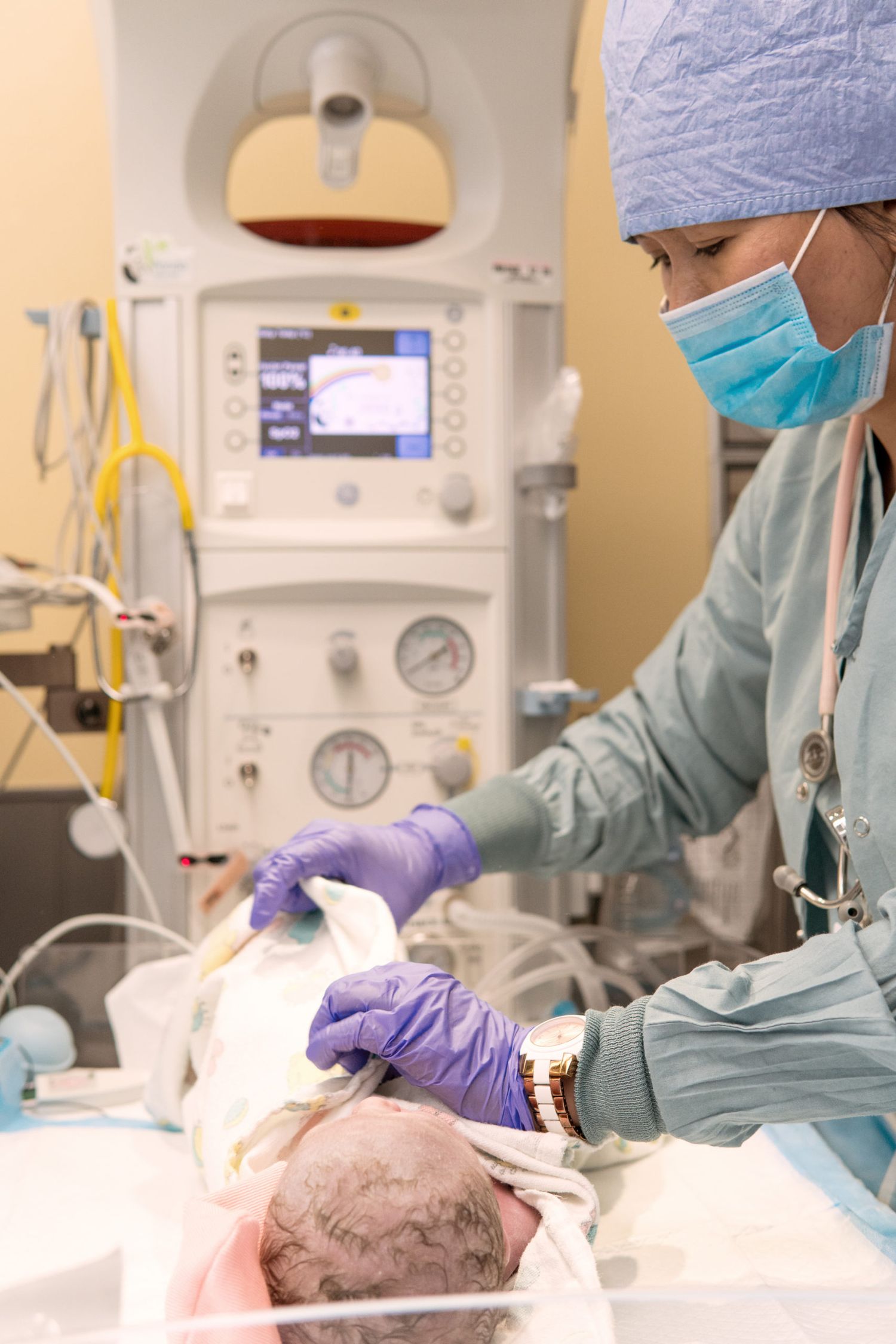 Healthcare worker wearing scrubs and mask attending to a patient on an examination table in a medical facility.