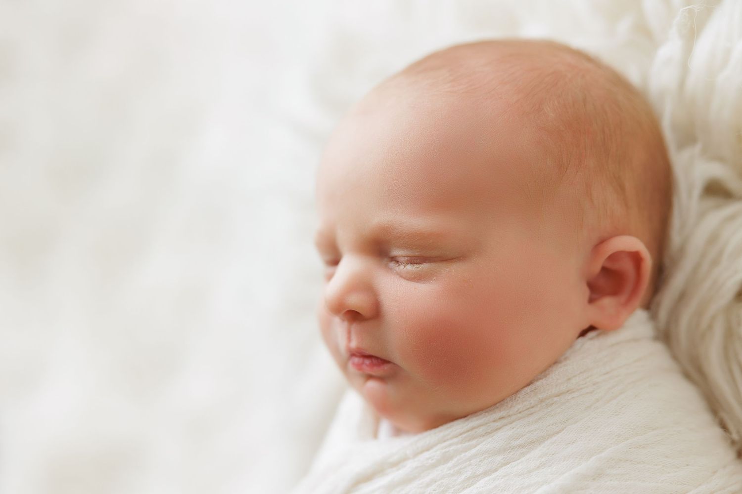 A sleeping newborn baby wrapped in white blankets rests peacefully against soft bedding.