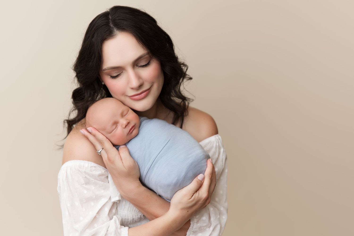 Woman in white lace dress holding baby against cream colored backdrop.