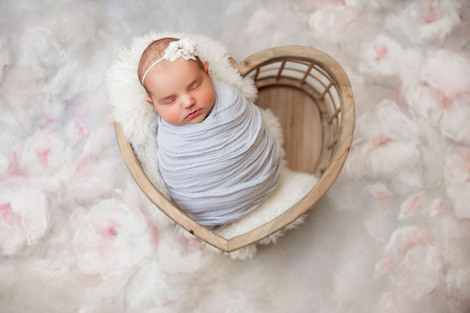 Baby swaddled in gray wrap sleeping peacefully in heart-shaped basket.