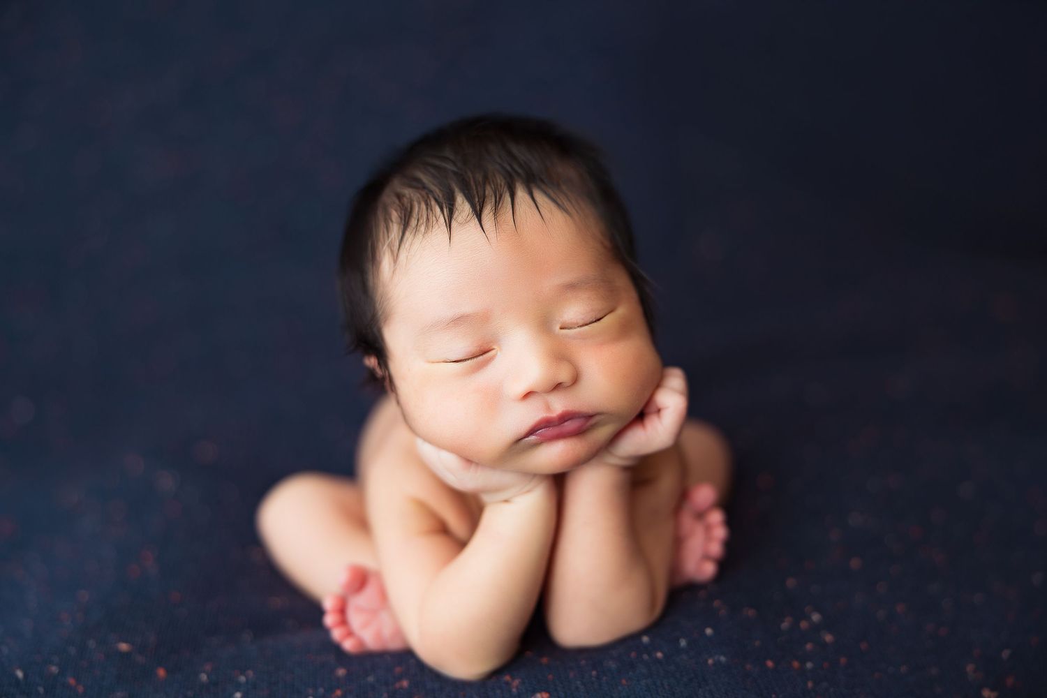 Sleeping newborn posed with hands under chin against dark background.