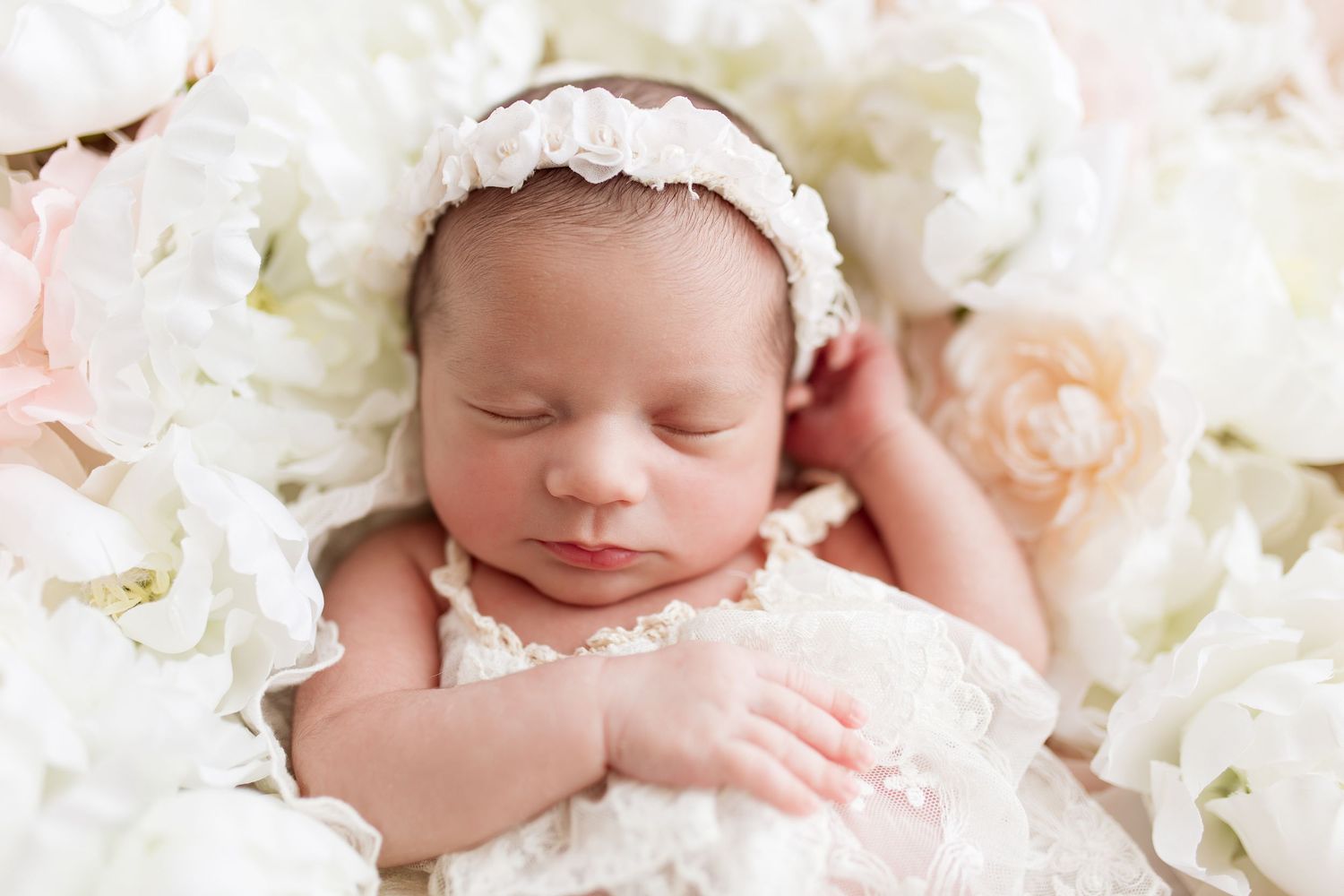 Peaceful newborn wearing white headband nestled among white roses.