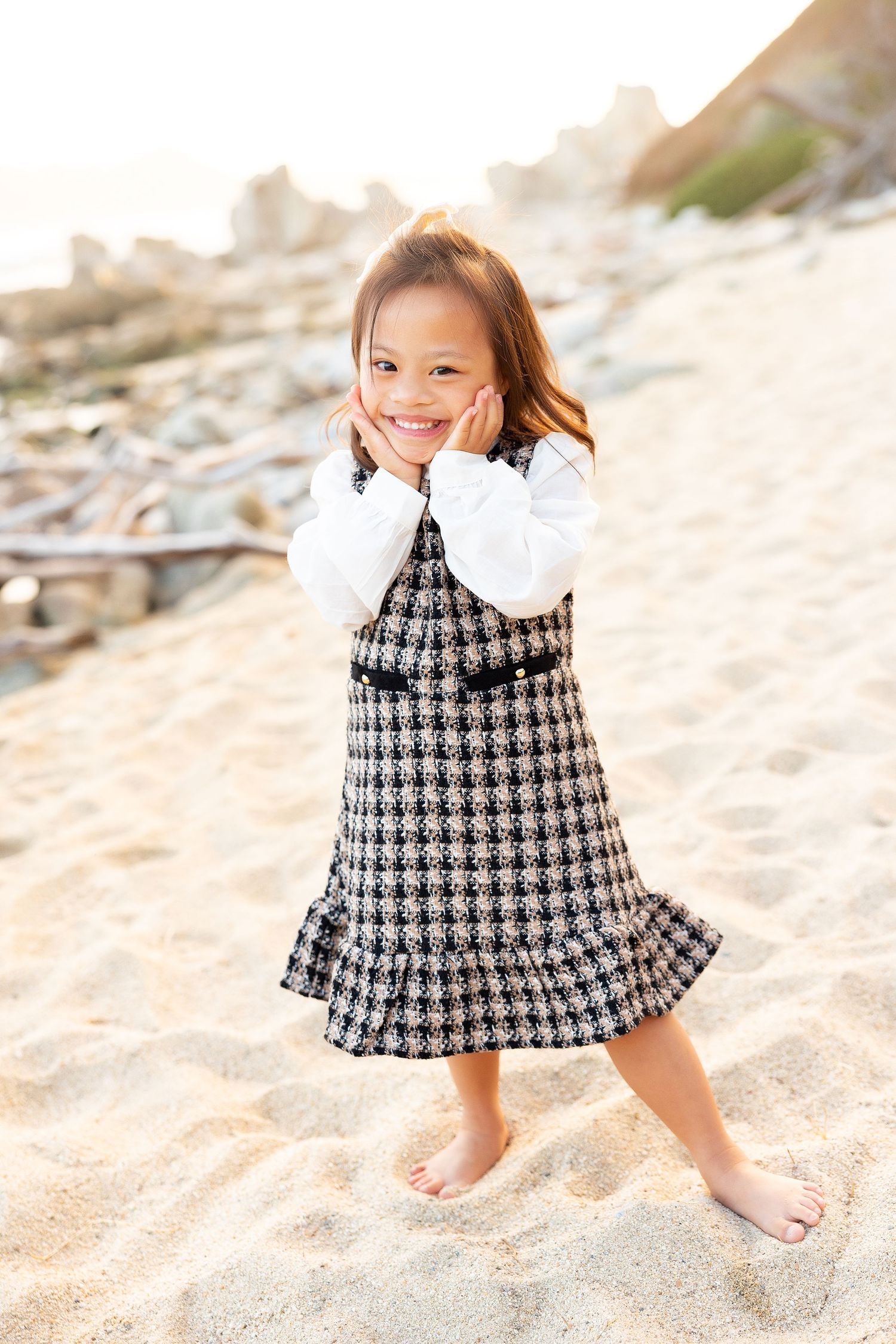 Child in houndstooth dress twirls playfully on sandy beach with cliffs in background.