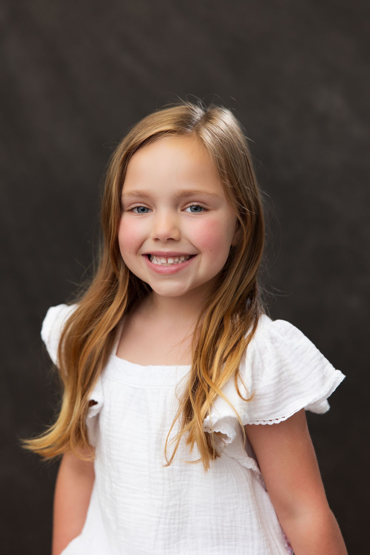 Young blonde girl wearing a white ruffled dress smiles warmly against a dark background in a studio portrait.