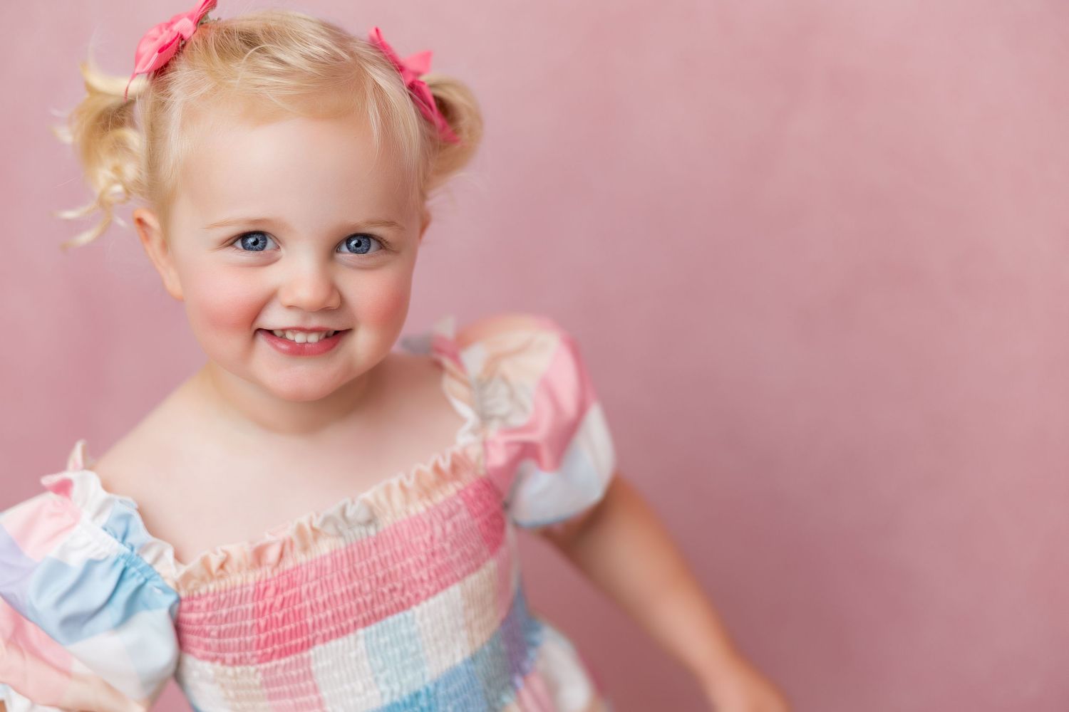 Sweet young toddler wearing pink and blue dress with hair bows poses against pink background.