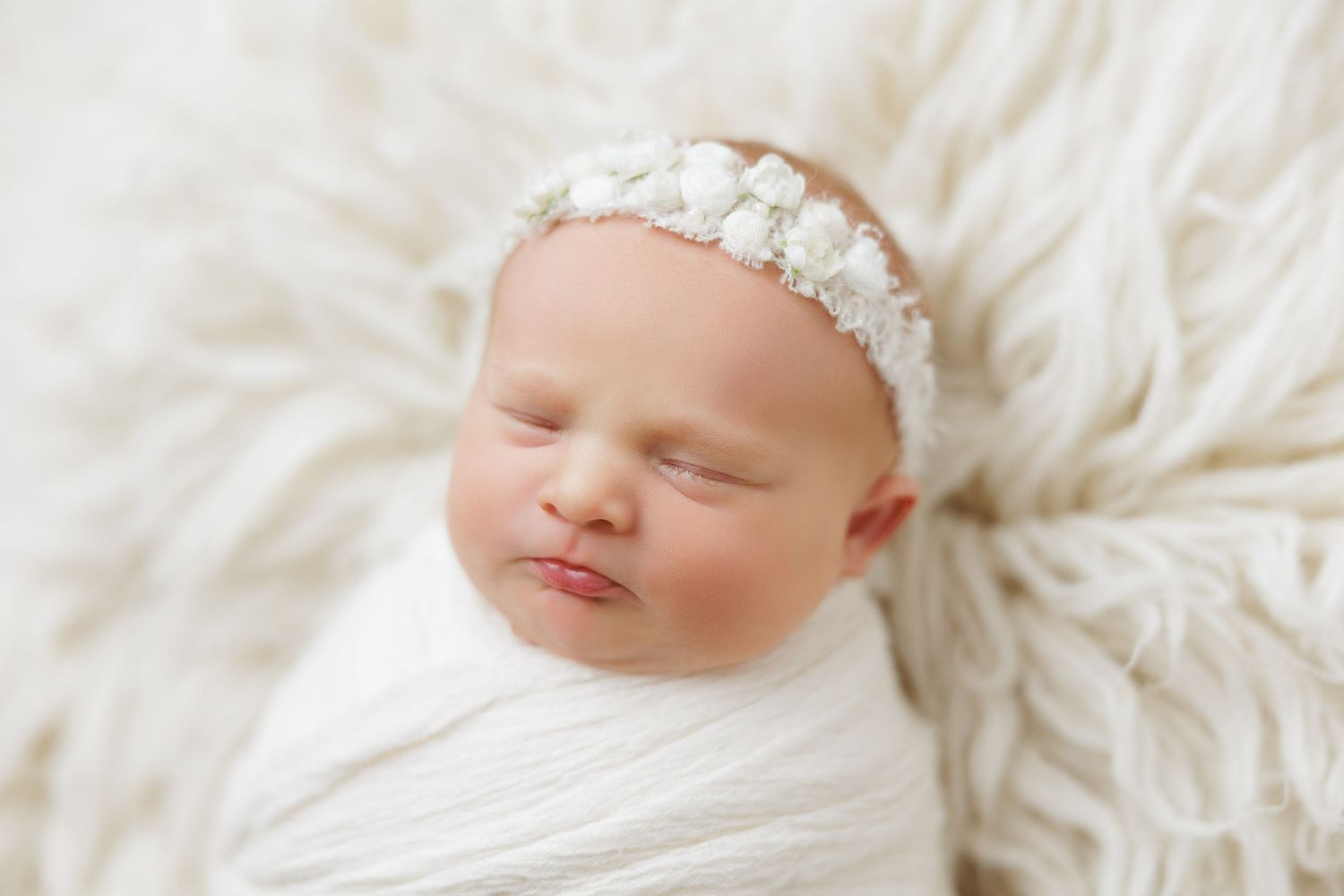 Sleeping newborn baby wrapped in white swaddle with floral headband on fluffy white fur backdrop.