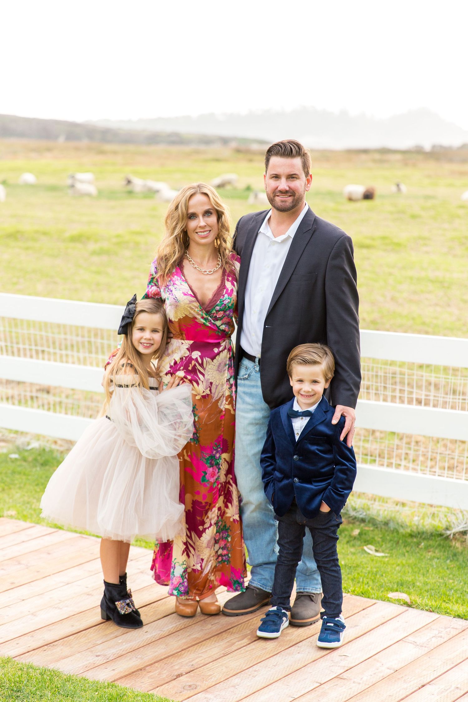 Family of four poses together by white fence on rustic farm with sheep in background.