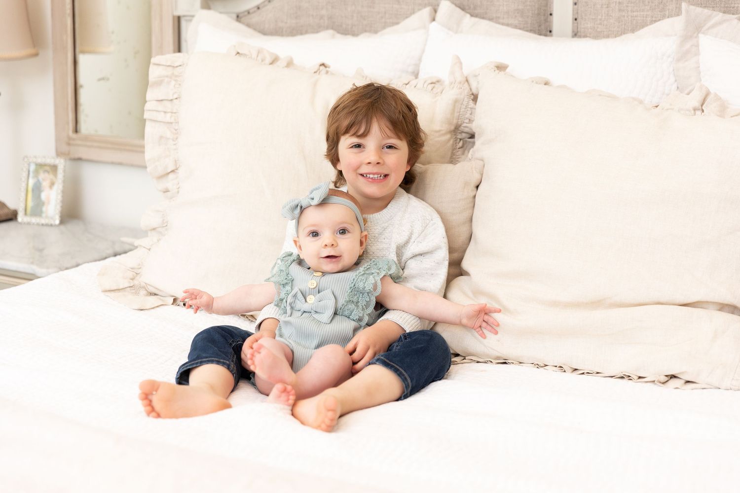 Two siblings sit together on white bedding in casual clothing.