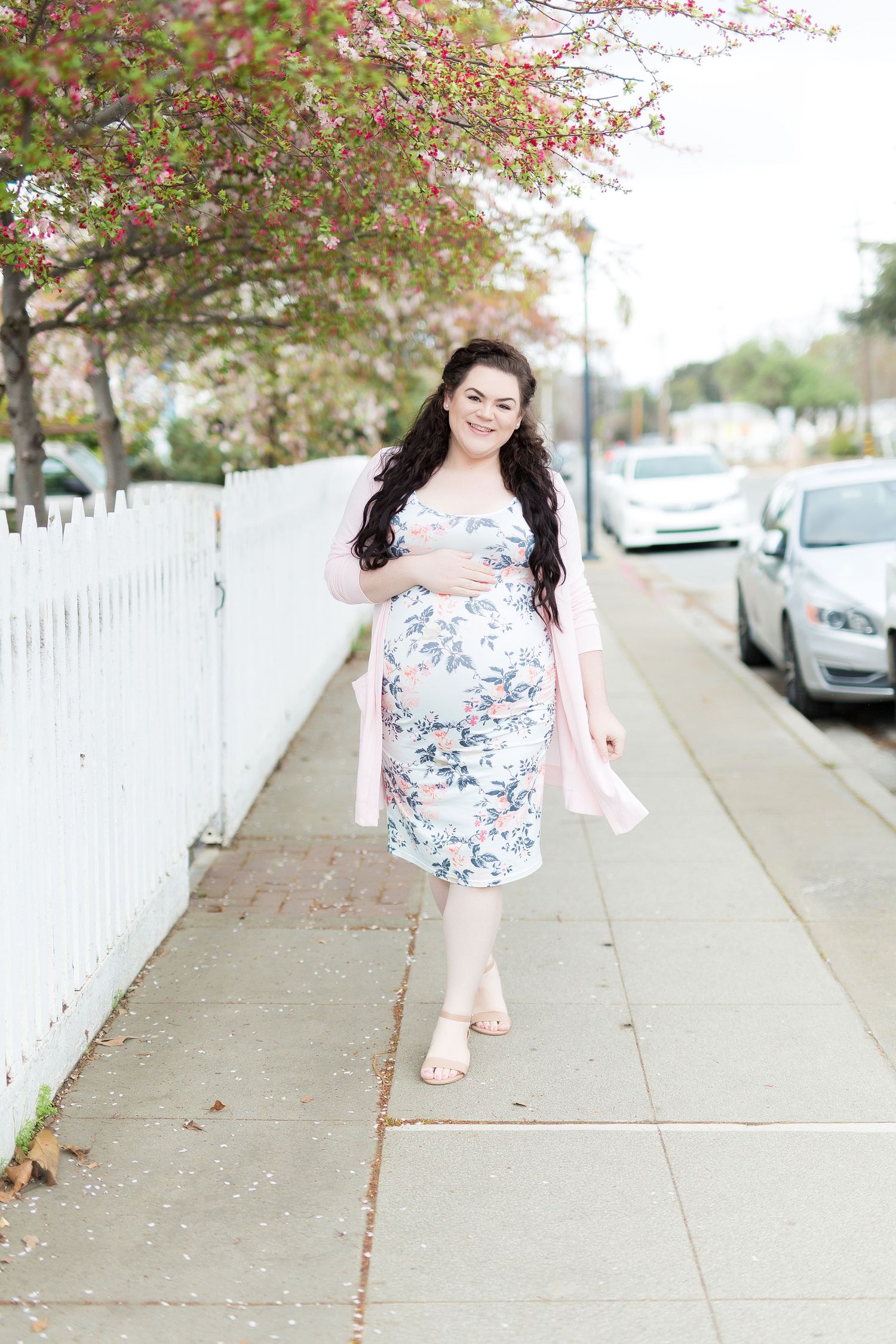 A floral dress and pink cardigan outfit styled for spring with a white picket fence and blooming trees in background, maternity.
