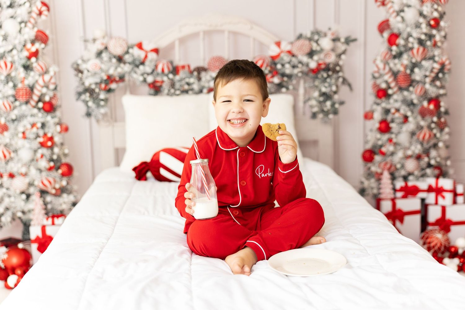 Child in red holiday pajamas sits on white bed with festive Christmas decorations and wrapped gifts in background.