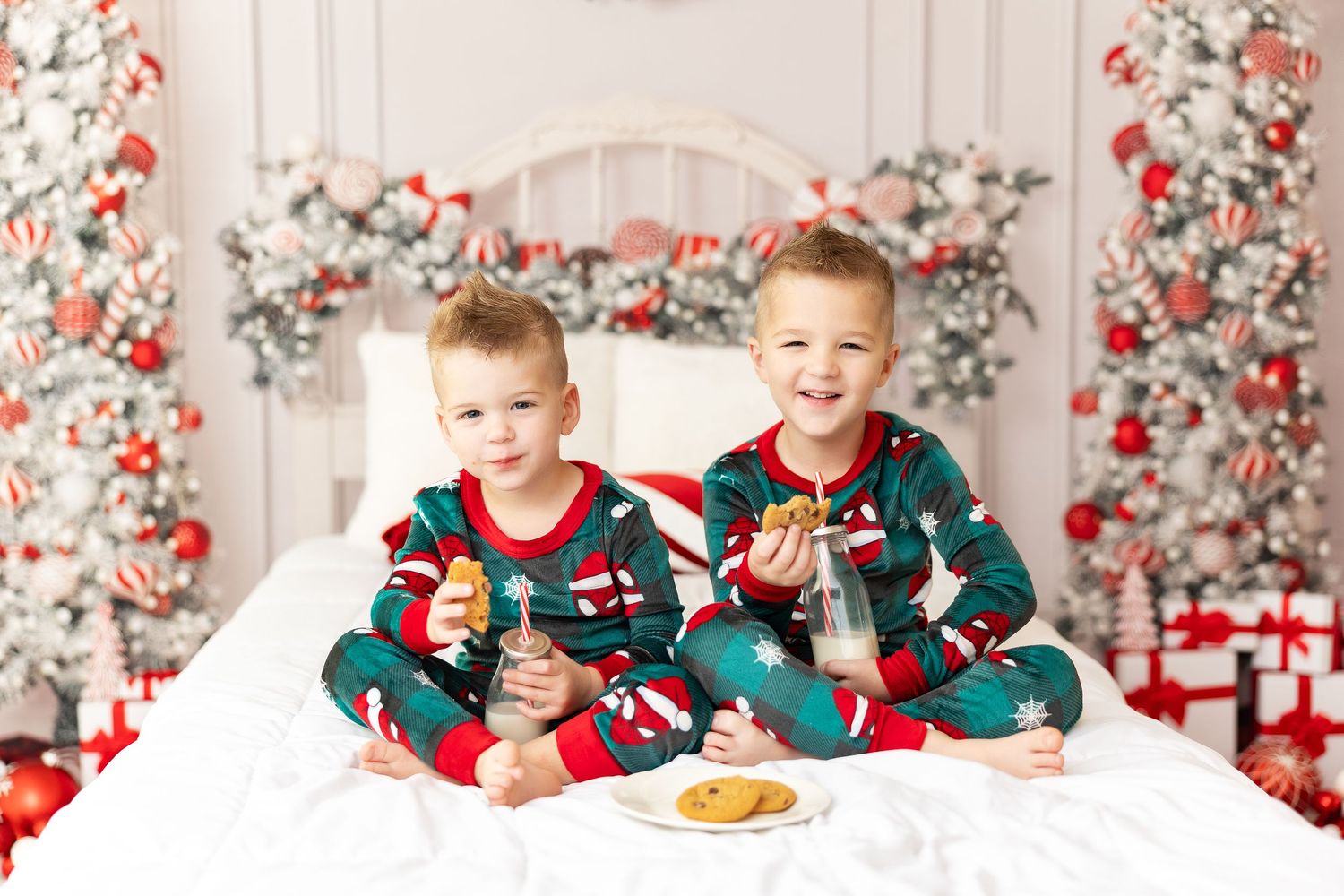 Two children in matching holiday pajamas enjoy cookies on a white bed with festive red and white decorations behind them.