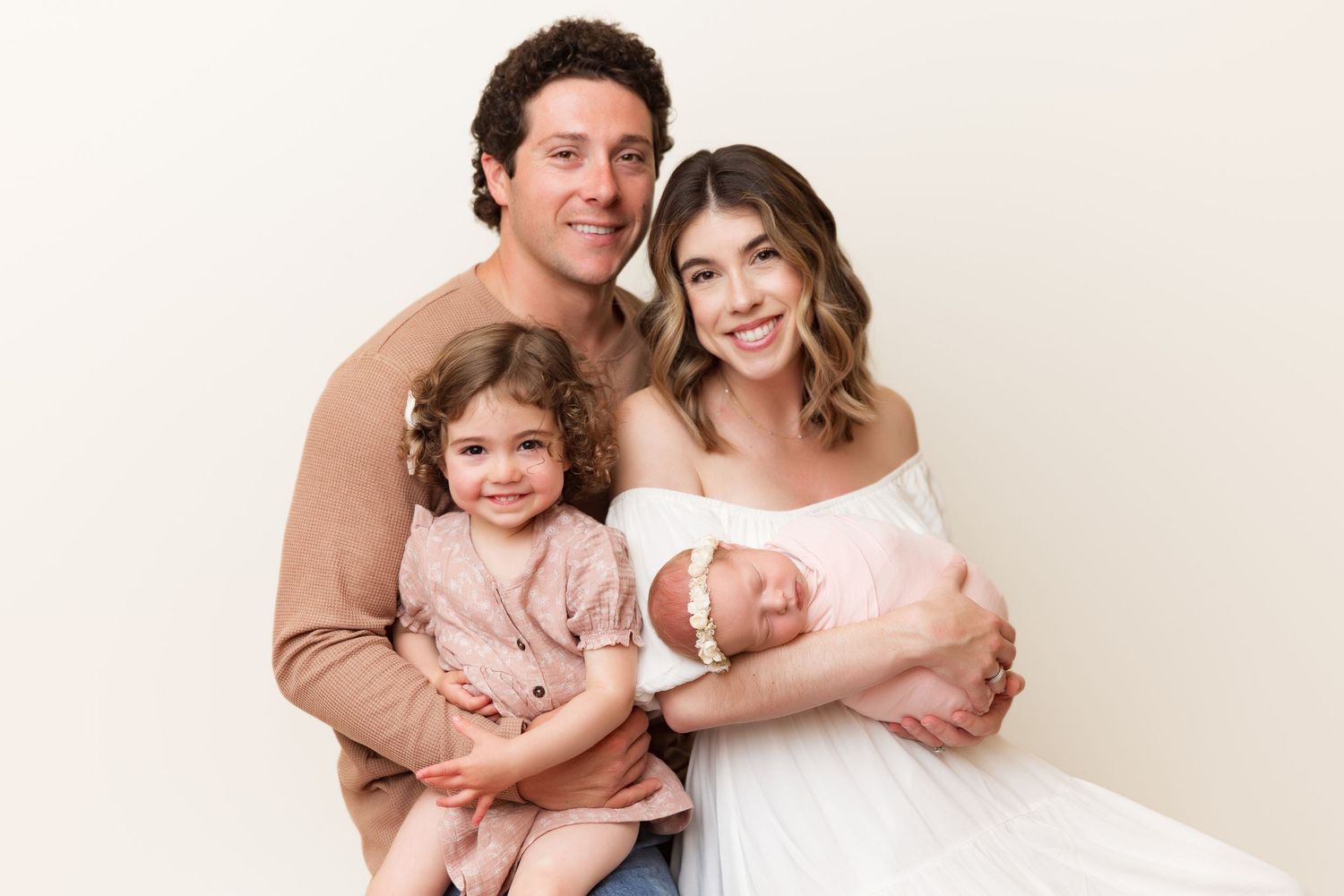 Studio portrait showing a family of three in soft neutral tones with a newborn baby and toddler.