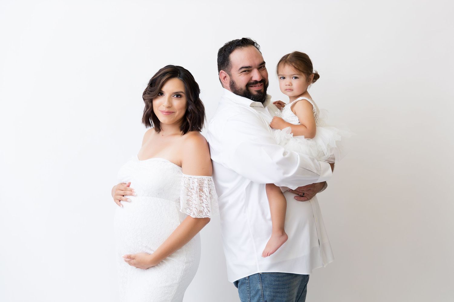 Family maternity photoshoot with parents and toddler in white clothing against white background.