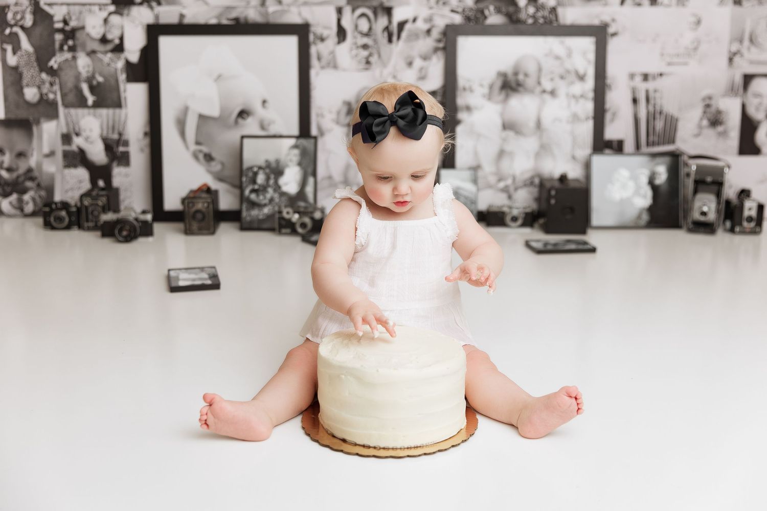 Baby in white outfit and black bow sitting with birthday cake against photo wall backdrop.