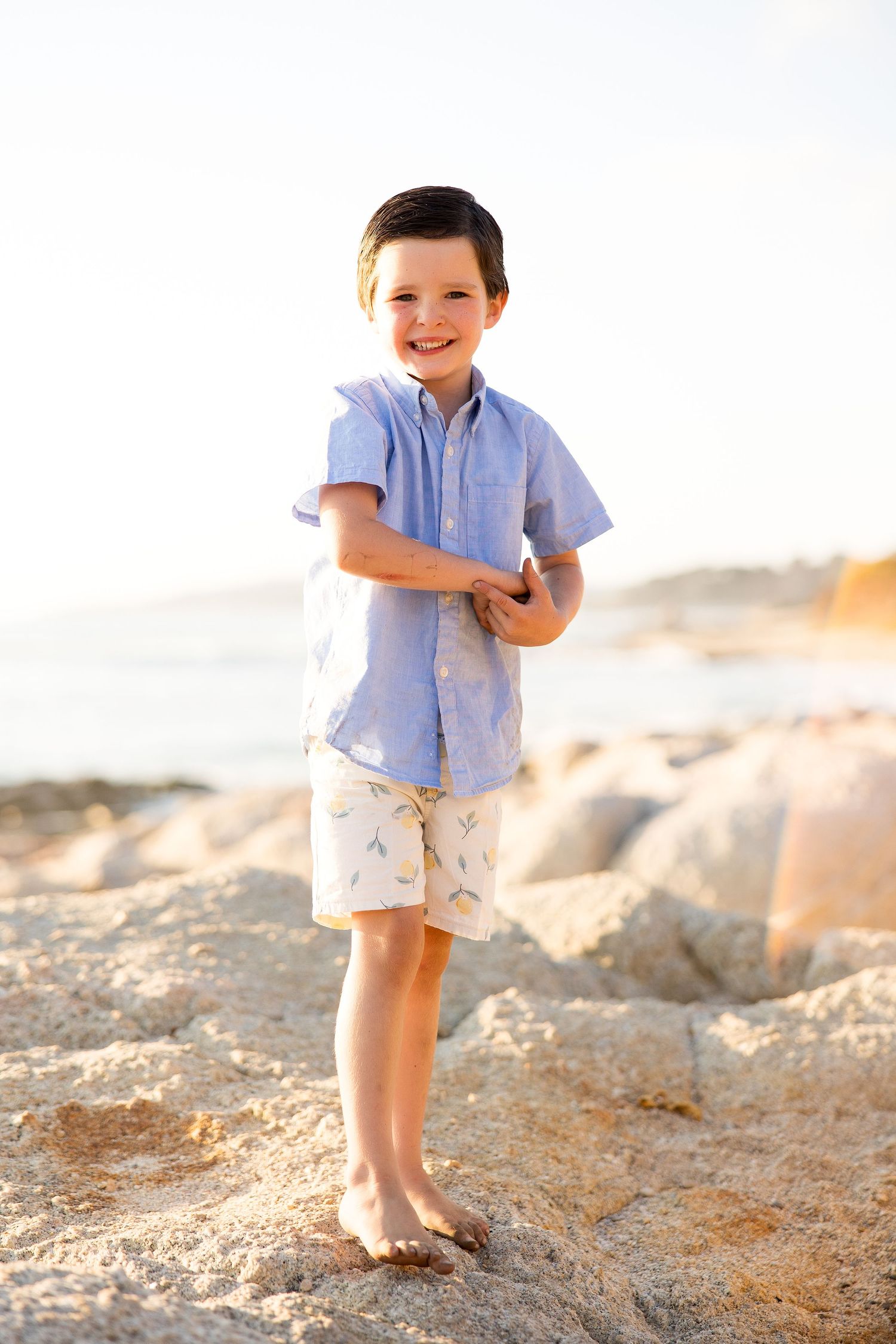 Child in light blue shirt and khaki shorts standing barefoot on rocky beach at sunset.