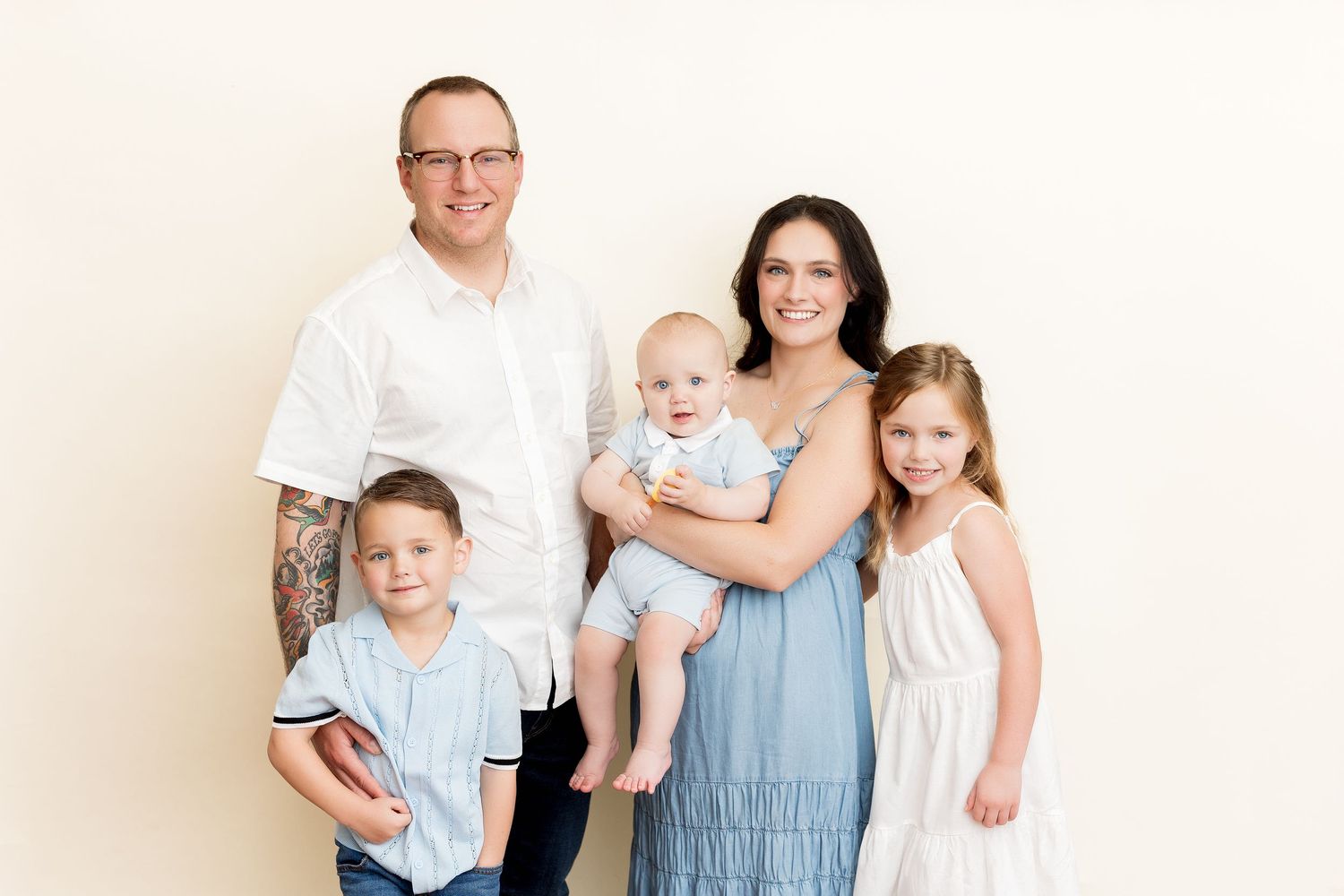 A family of five poses together against a cream colored background wearing coordinated blue and white outfits.