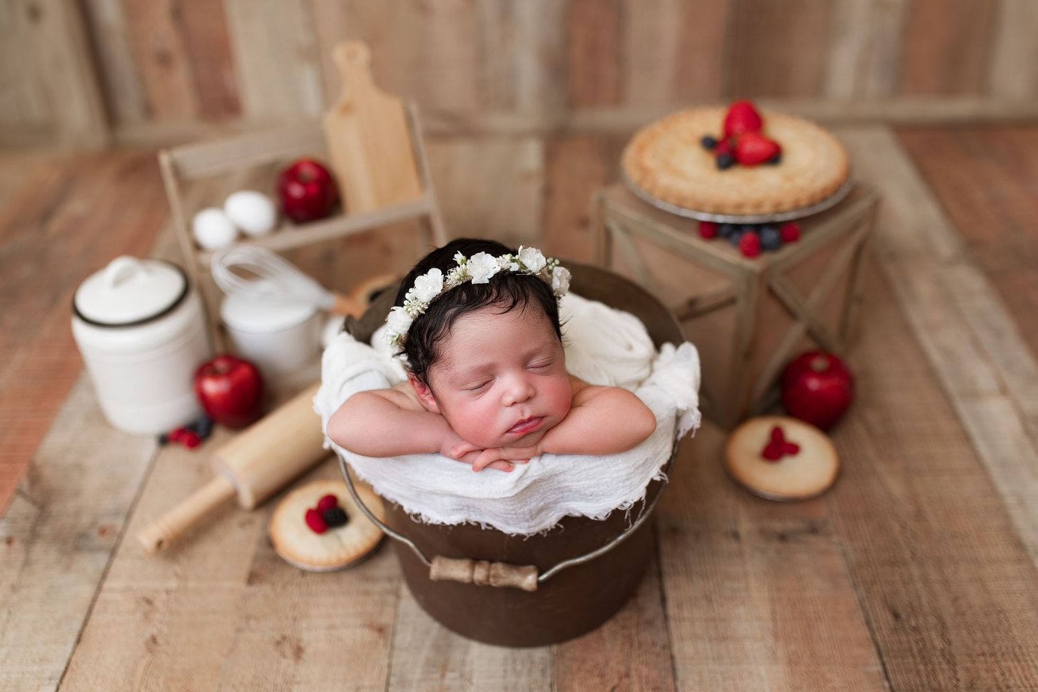 Newborn photography setup with wooden props, white fabrics, and red raspberries arranged on rustic wooden floor.