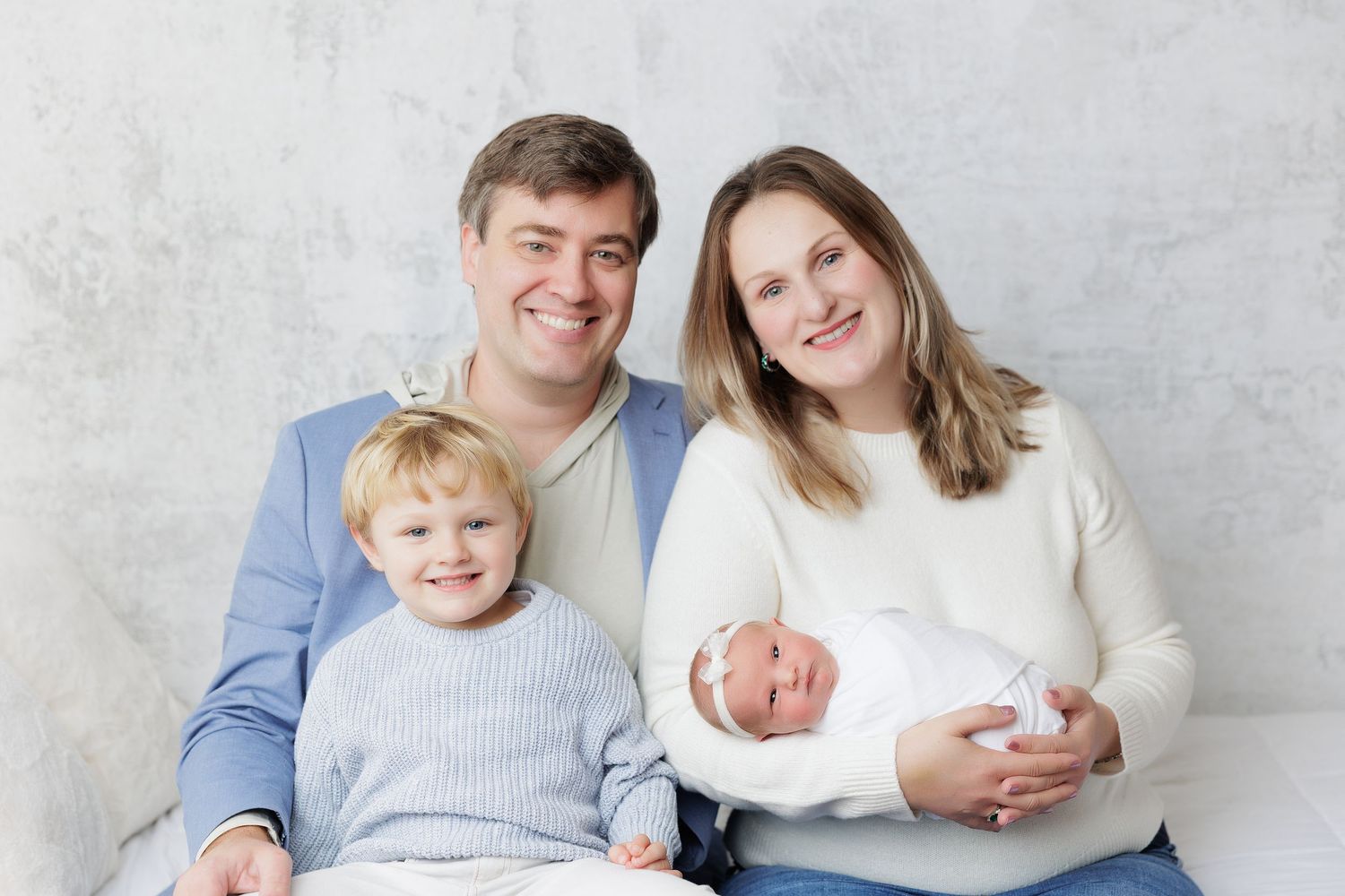 Family portrait with parents and two children, including a newborn baby, smiling together on a white couch.