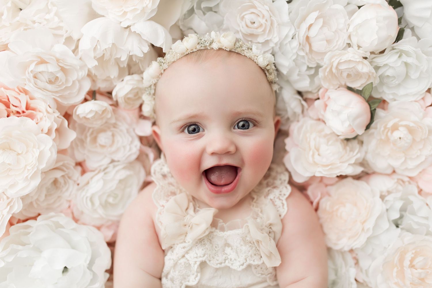 Baby in delicate lace dress surrounded by white and blush pink roses during portrait session.