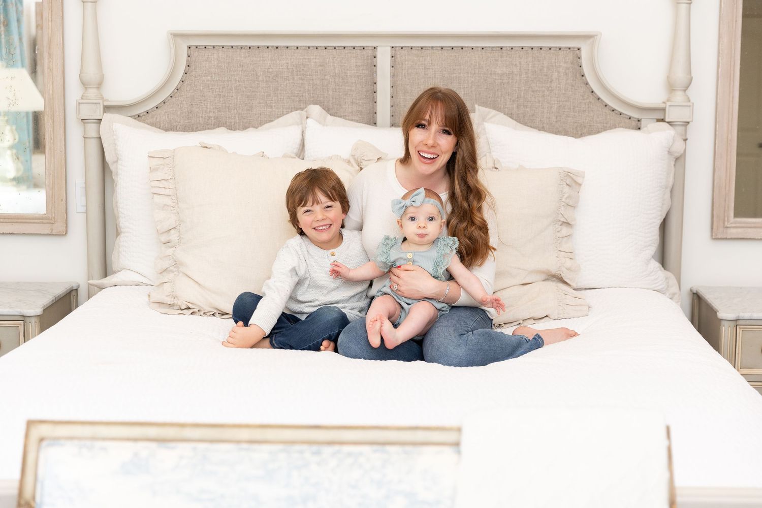Family lounging together on a plush white bed in a bright, airy bedroom setting.