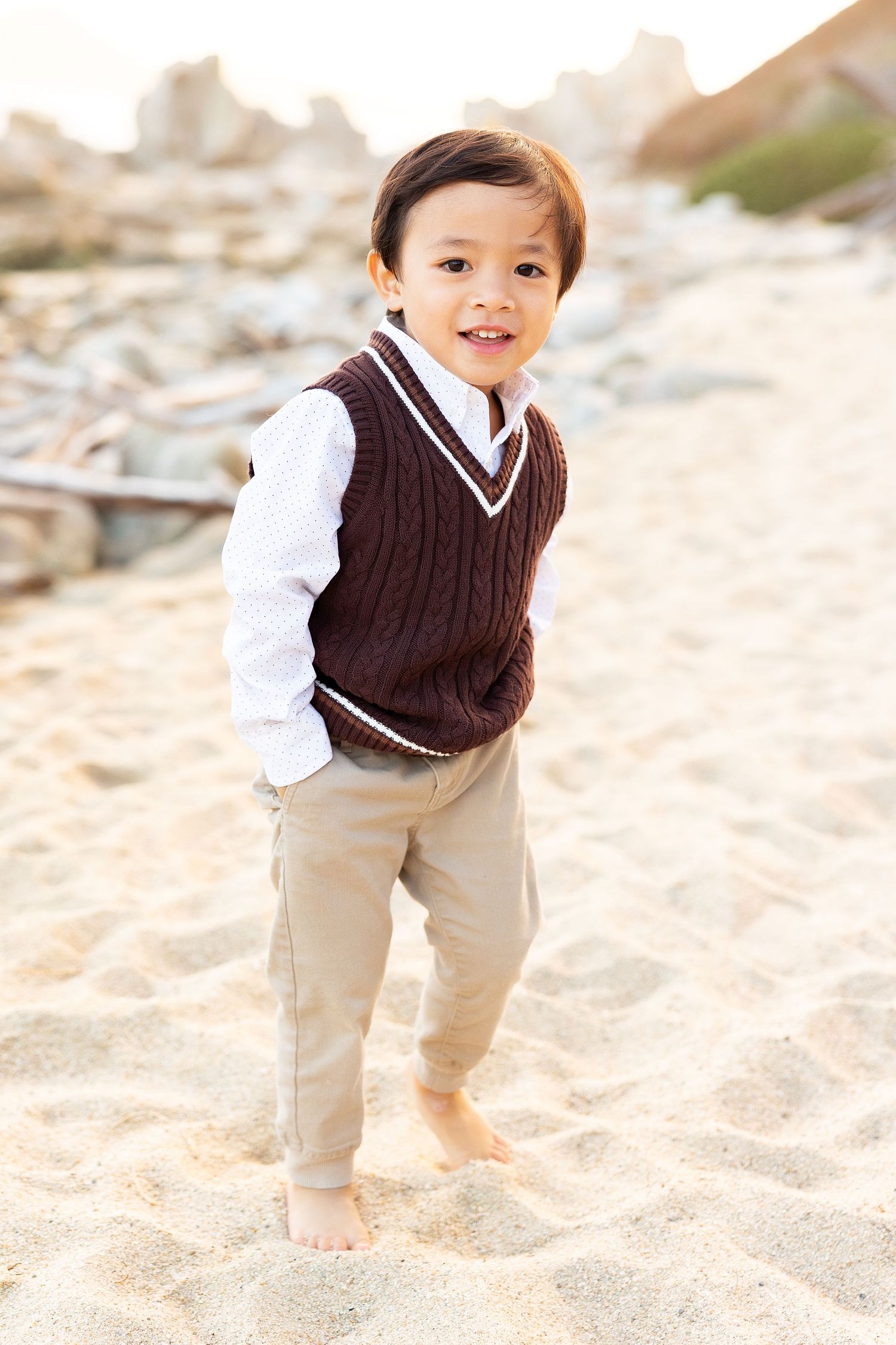Young child in preppy sweater vest outfit standing barefoot in beach sand.