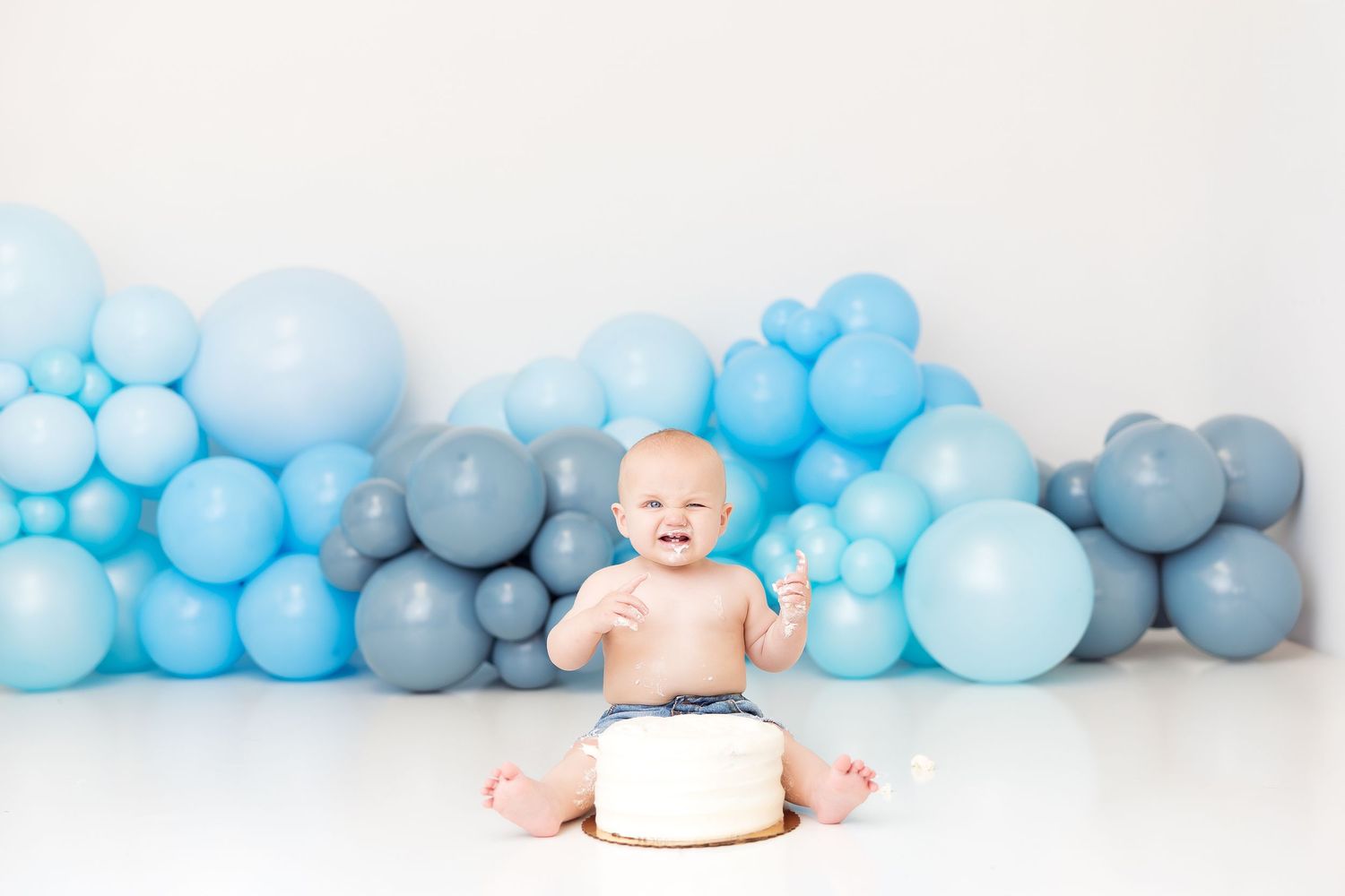 Baby boy sits smiling in front of blue balloon wall with white smash cake for first birthday photo.
