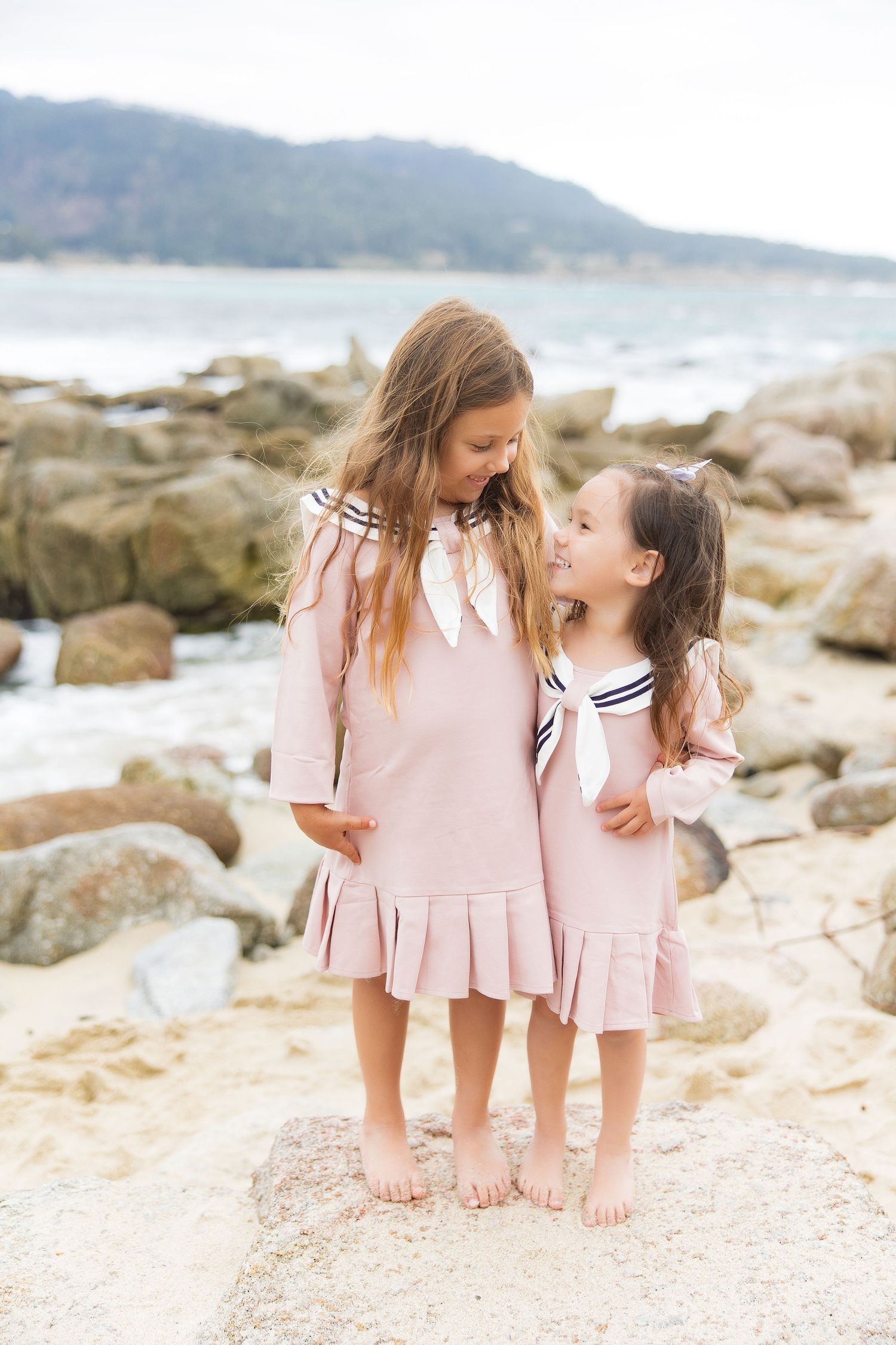 Two young girls in matching pink dresses with sailor scarves smile at each other on a Carmel River Beach.
