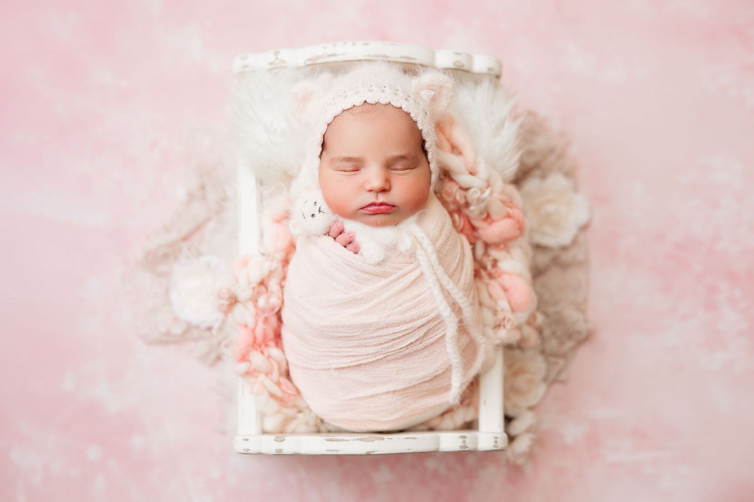 Sleeping newborn wrapped in cream fabric on pink textured backdrop with floral accents.