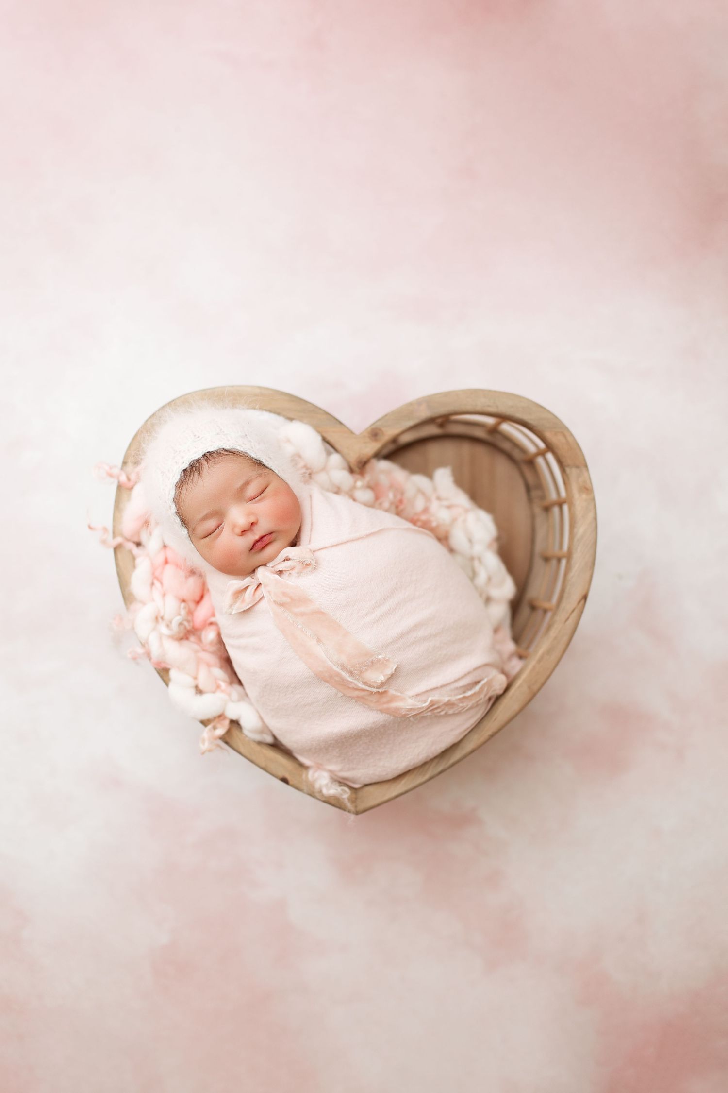 A sleeping newborn baby swaddled in pink lies peacefully in a heart-shaped wooden basket on a soft white background.