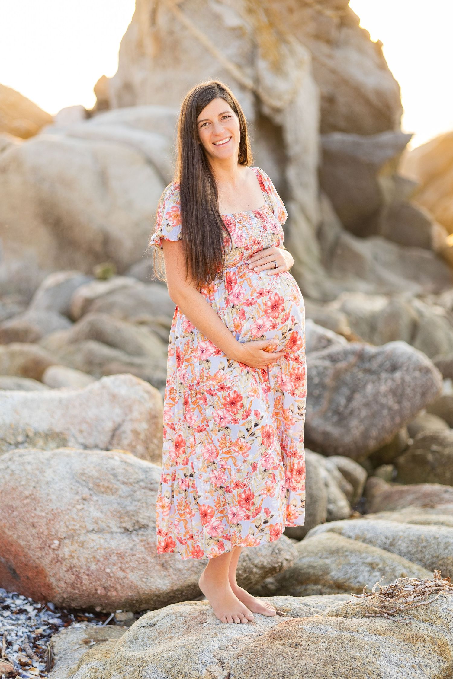 Woman in floral dress poses gracefully on coastal rocks during sunset photography session.