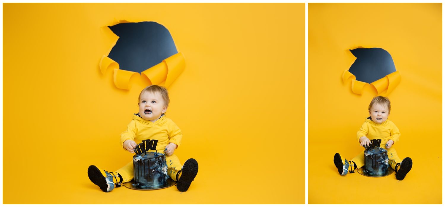 A baby in a blue graduation cap and gown sits and plays against a bright yellow background.