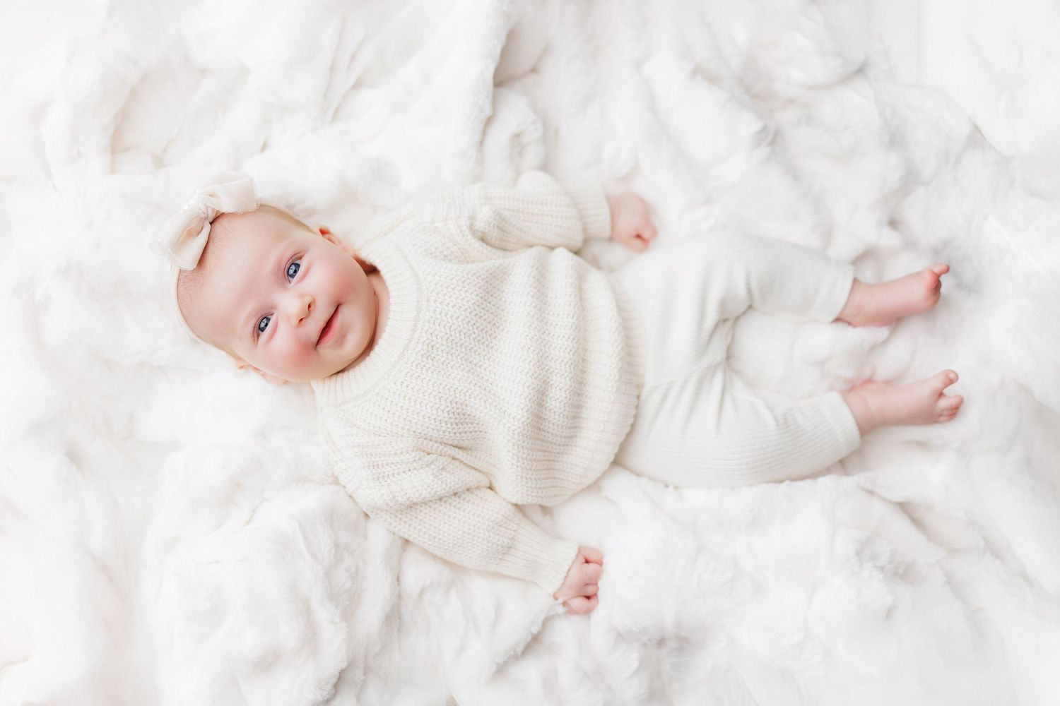 A smiling baby wearing a white knit sweater lies on soft white blankets.