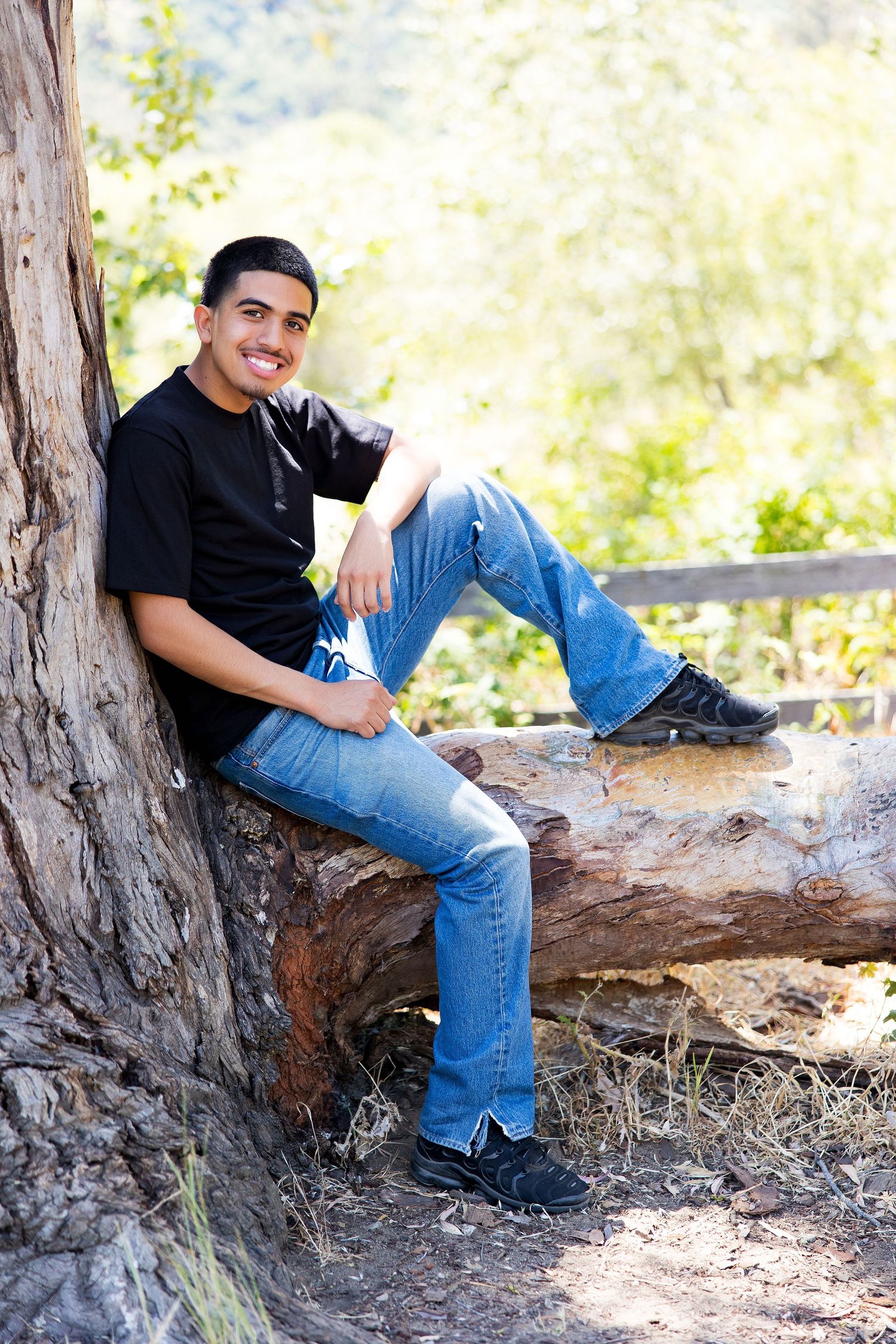 Young man in black shirt and jeans sitting on a log leaning against a tree outdoors smiling.