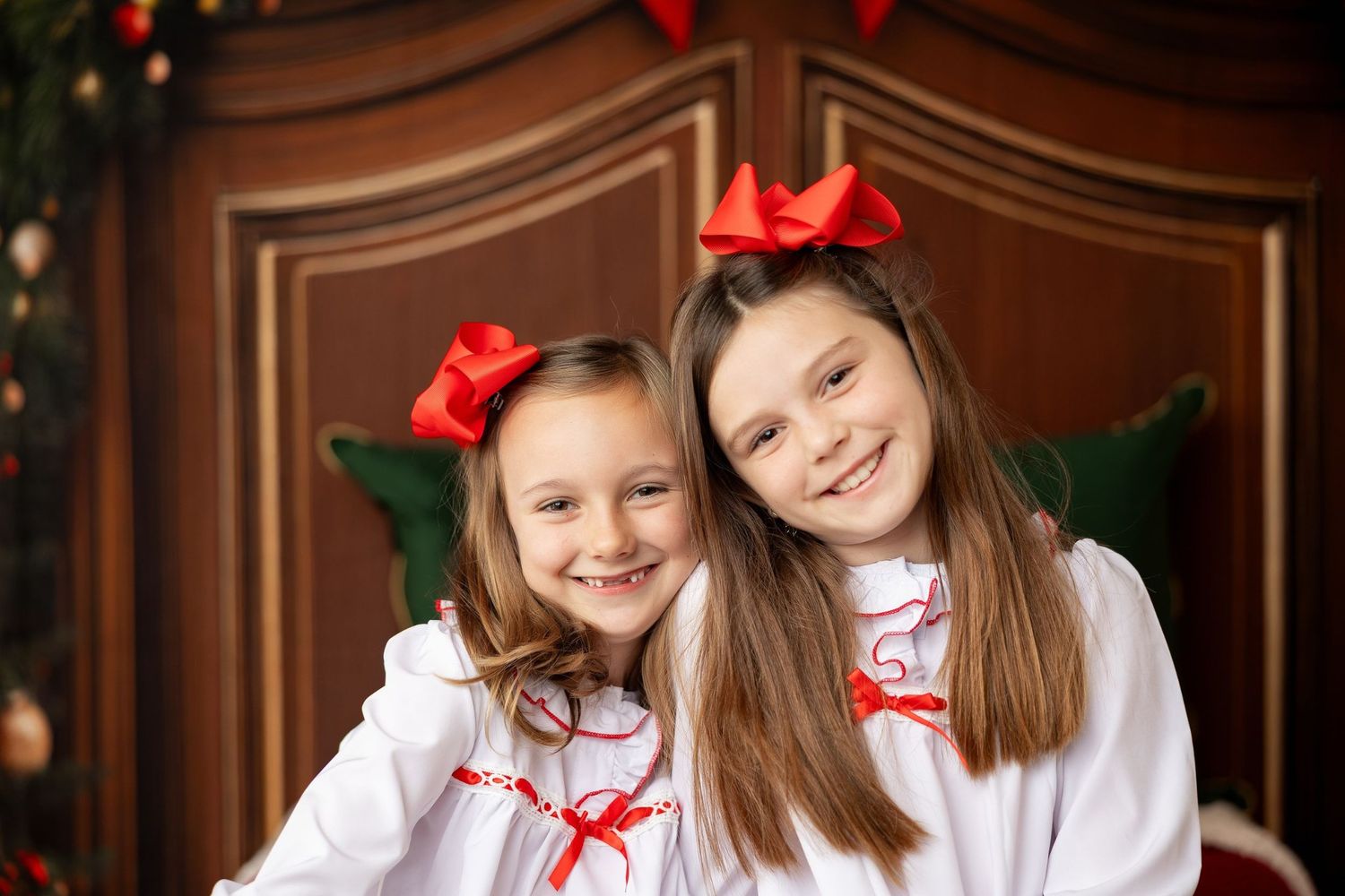 Sisters in white holiday outfits with red bows by the Christmas tree at a Milk & Cookies mini session in Pacific Grove