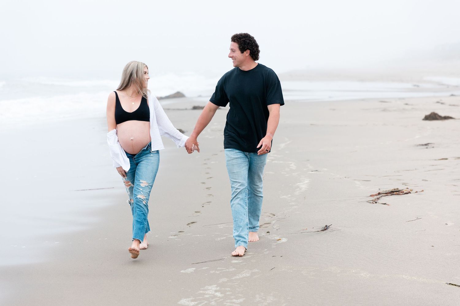 maternity couple mom wearing a black bra, white shirt and jeans at the beach