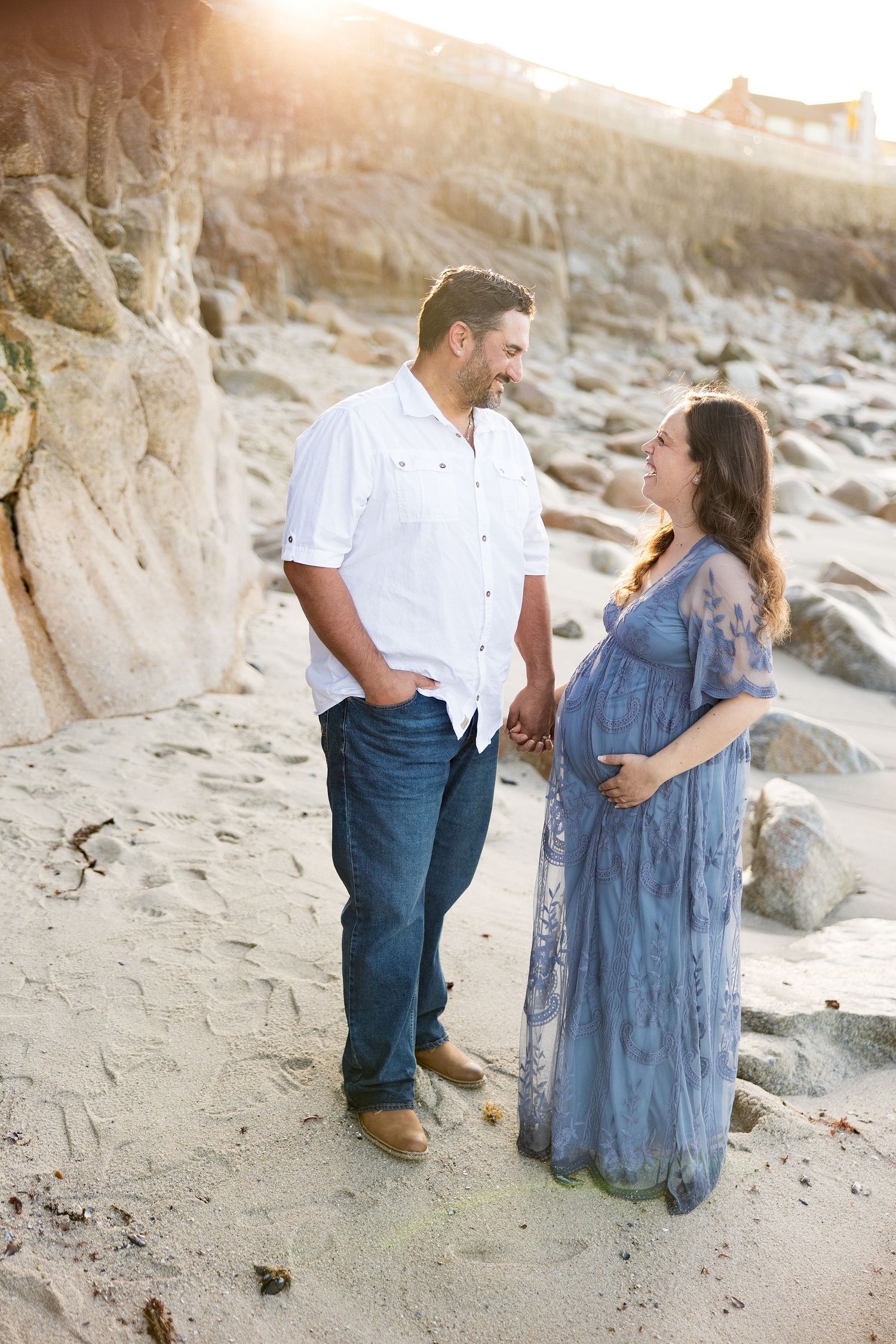 Pregnant couple holding hands at Lover's Point man in white shirt and jeans, woman in flowing blue maternity dress, golden sunset.