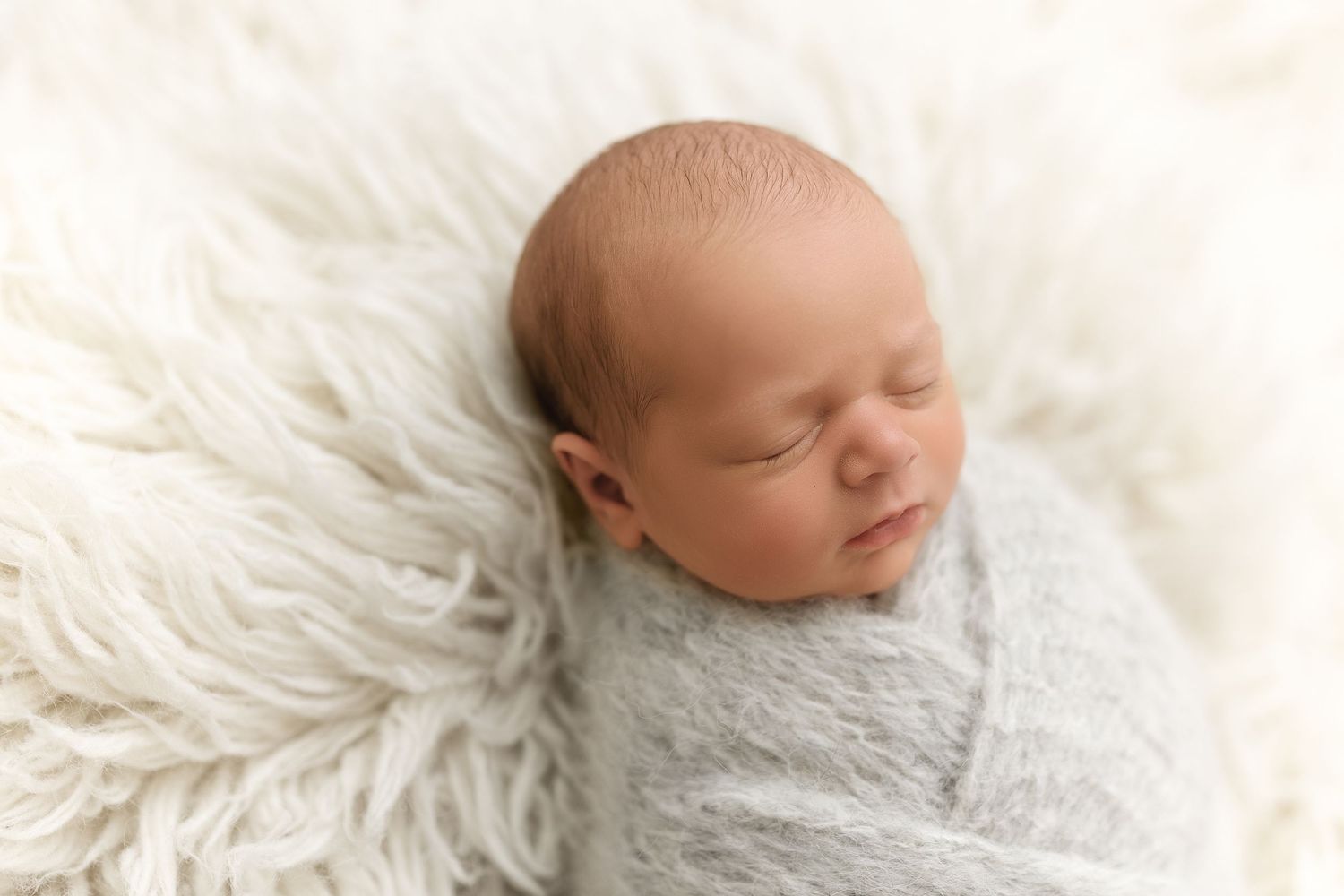 Peaceful sleeping newborn wrapped in soft gray swaddle blanket resting on white fluffy fur backdrop.