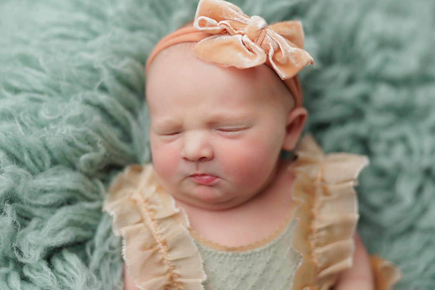 Newborn baby girl sleeping peacefully on teal fur, wearing a peach ruffle outfit and matching bow headband.
