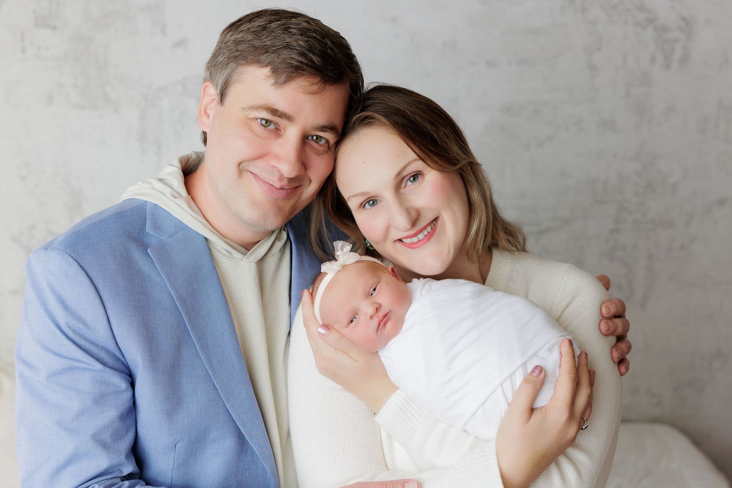 Happy parents smiling and holding their newborn baby wrapped in white, posing for a professional family portrait.